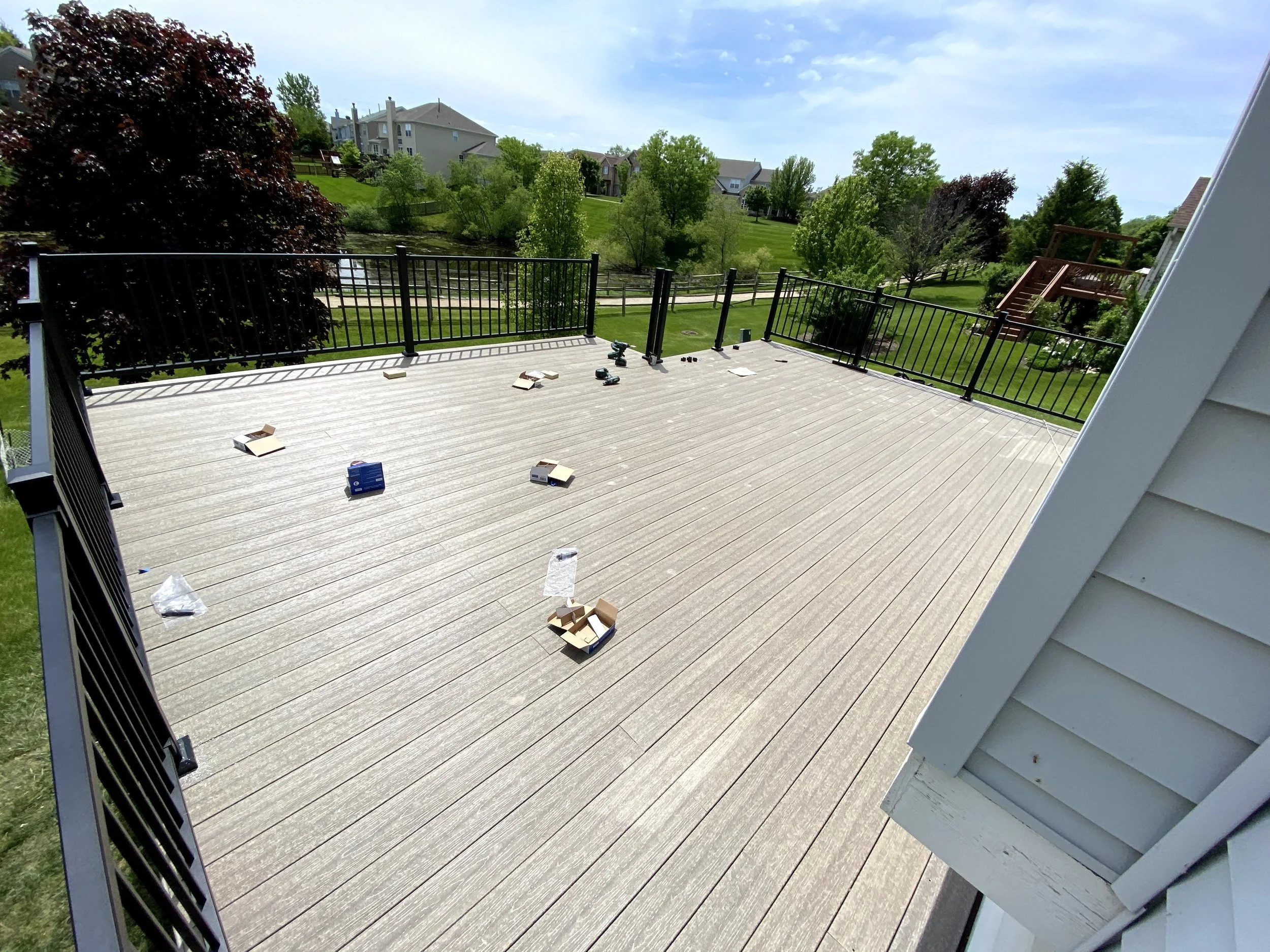 Empty outdoor deck with some open boxes and scattered items, overlooking a green backyard with trees and houses in the distance.