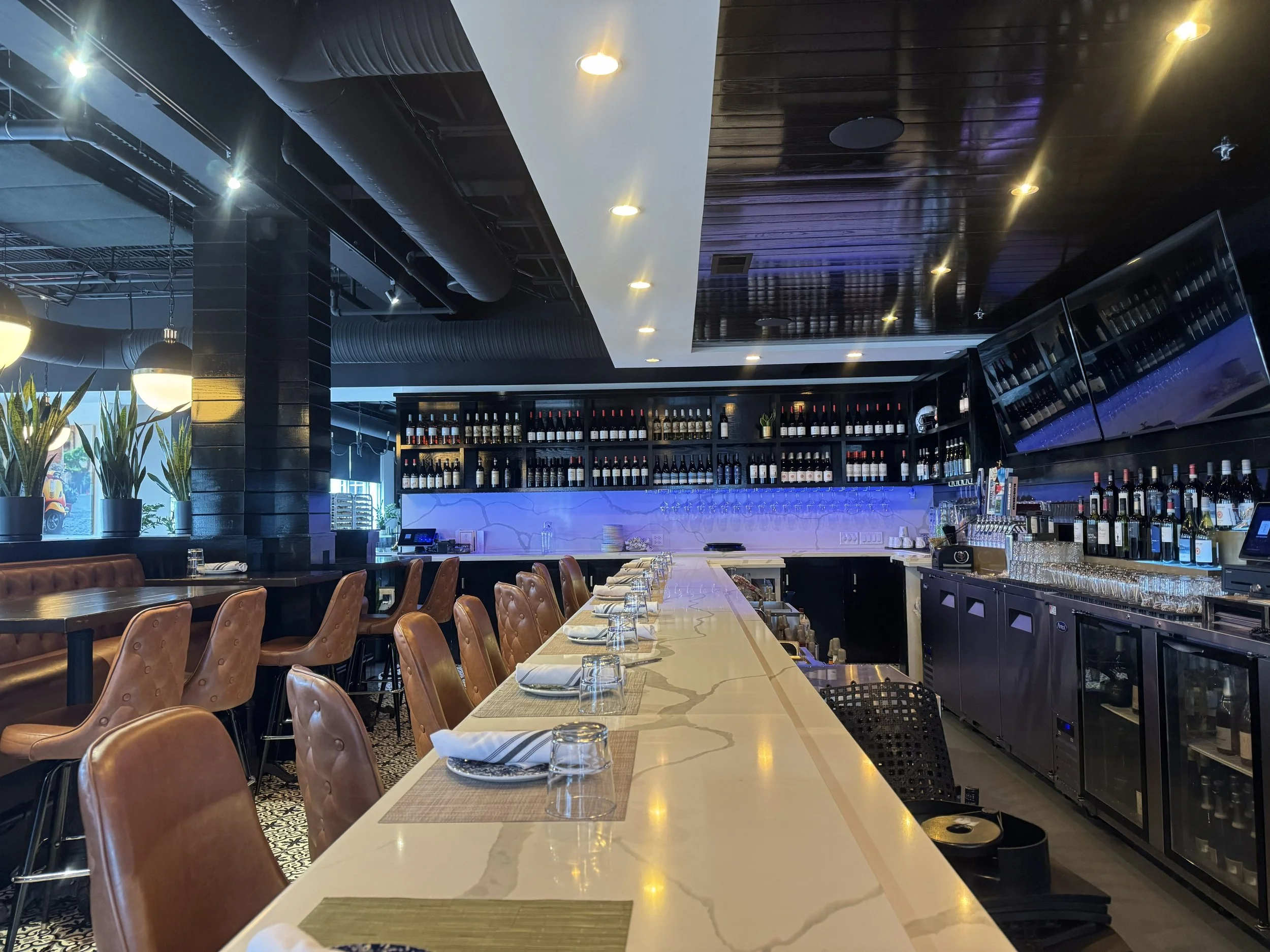 Interior of a modern bar with a long white marble counter, brown leather bar stools, bottles of wine and liquor on shelves, wine glasses hanging upside down, and multiple large TV screens on the wall.