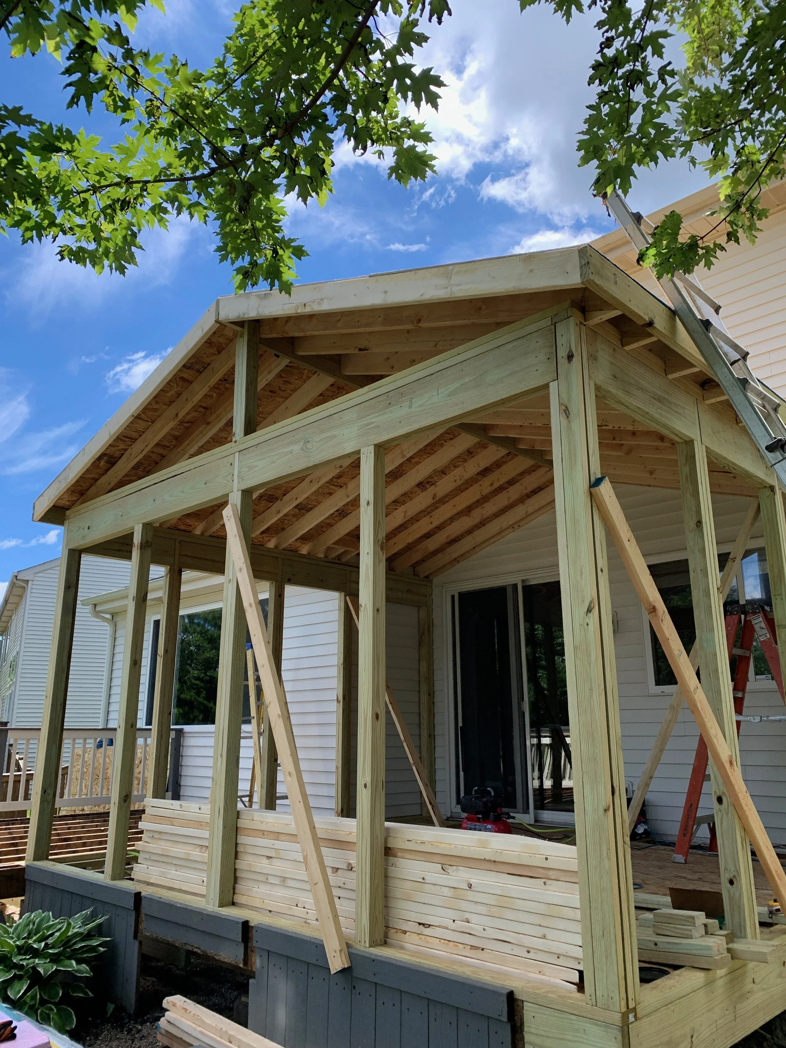 Construction of a wooden deck or porch extension on a house, with framing partially built and a ladder leaning against the structure, under a blue sky with some clouds and green tree leaves.
