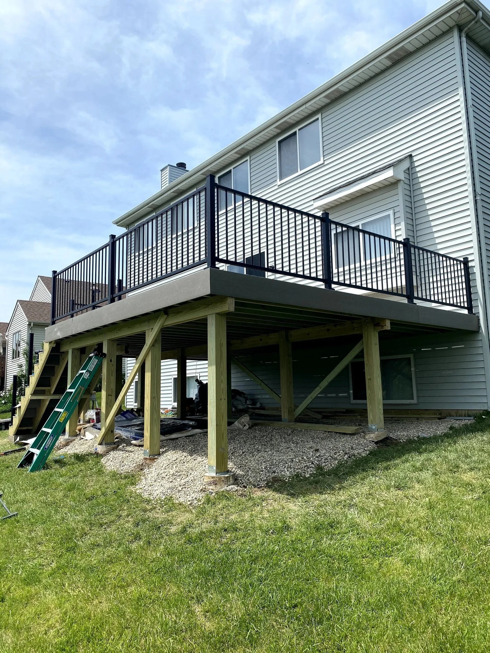 A house with a newly constructed elevated deck with black railing and stairs, supported by wooden beams, with construction tools and materials underneath.