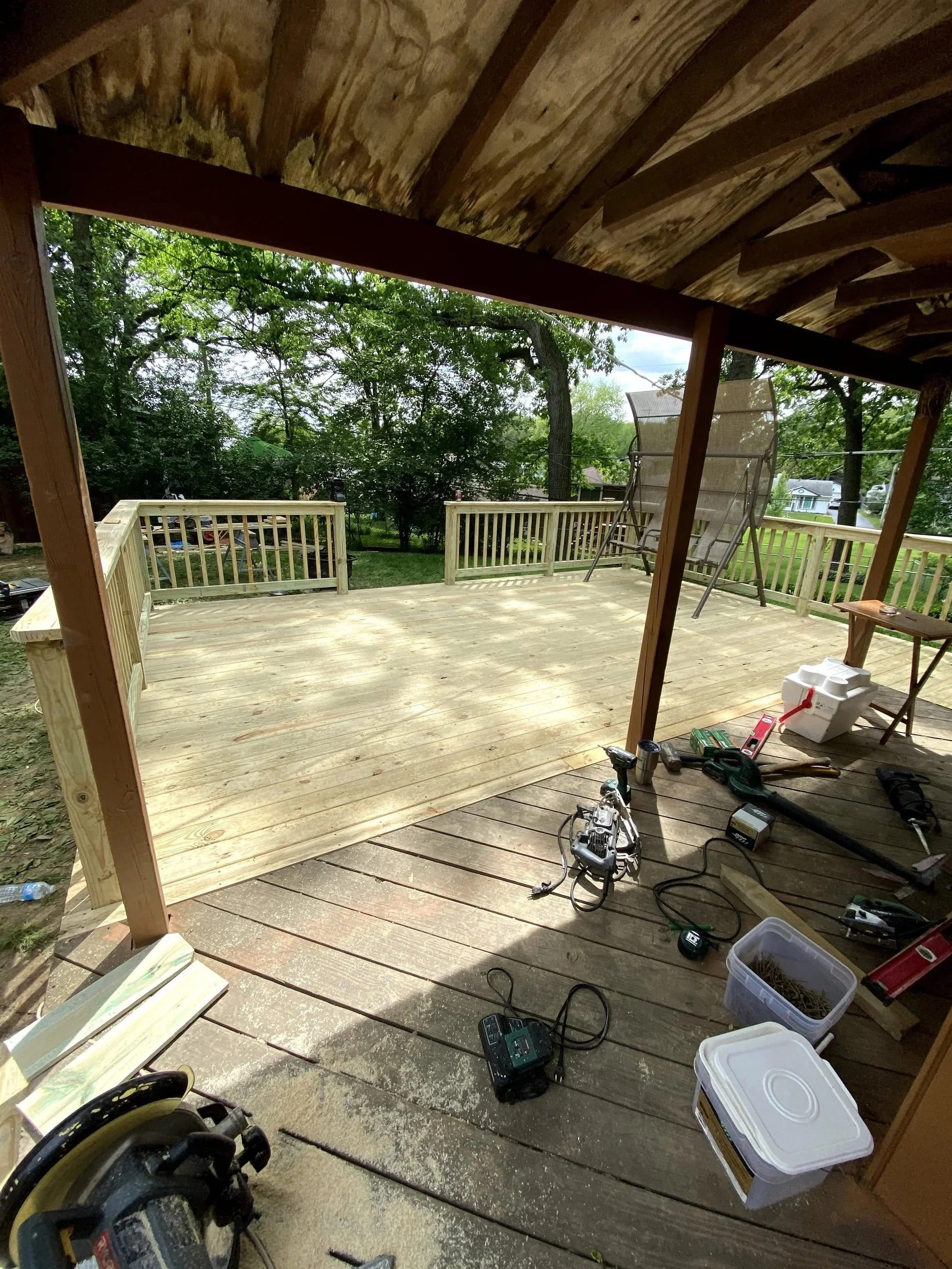 View of a wooden deck under construction with tools and materials scattered, surrounded by a green backyard with trees.