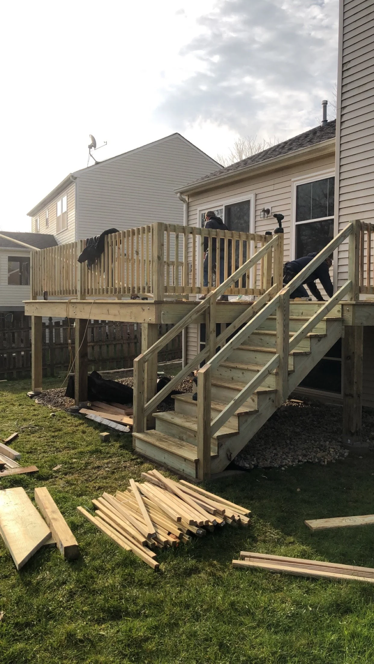 People working on building a wooden deck attached to a house, with stairs leading down to a grassy yard. Piles of wood and construction materials are on the ground.