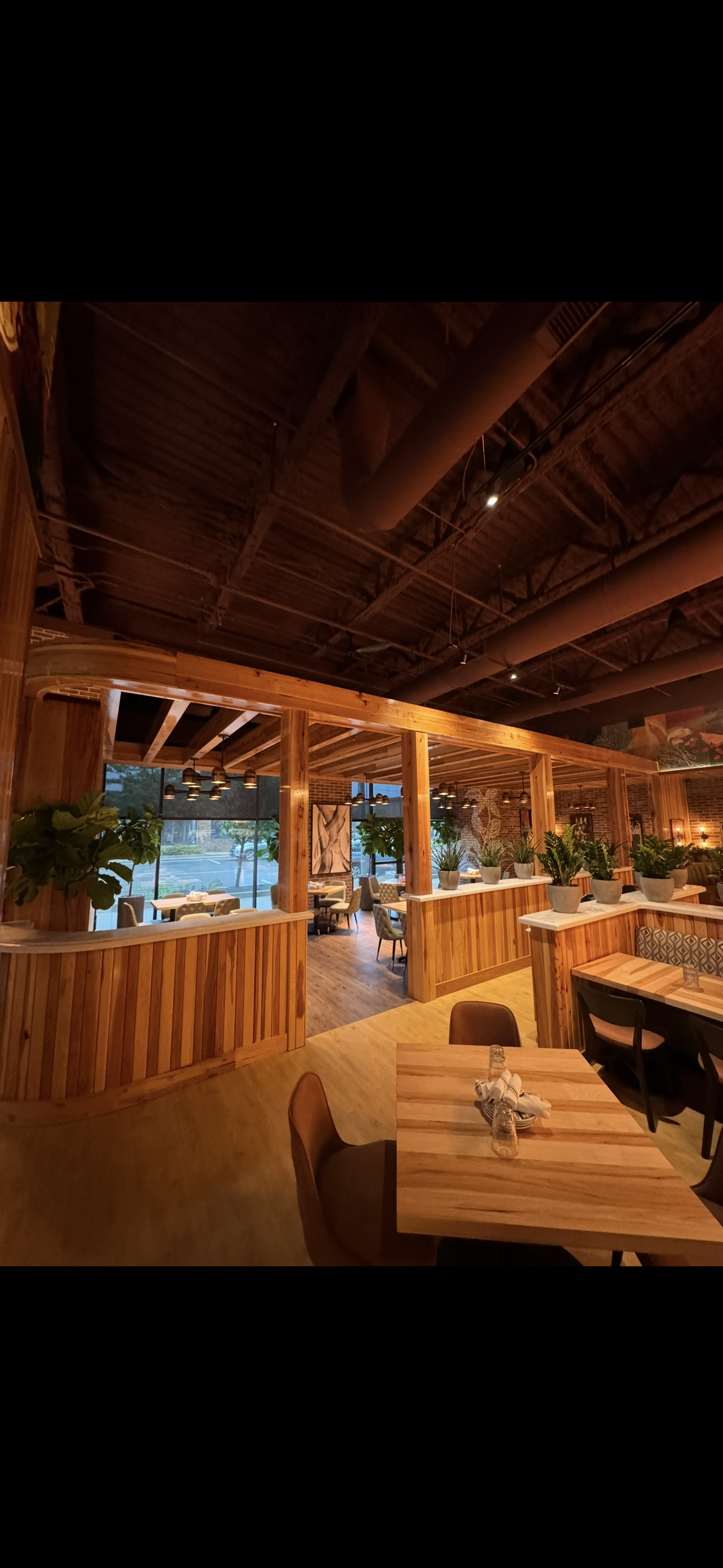Interior of a restaurant with wooden decor, plants, and seating areas with tables and chairs, lit by hanging pendant lights and large windows.