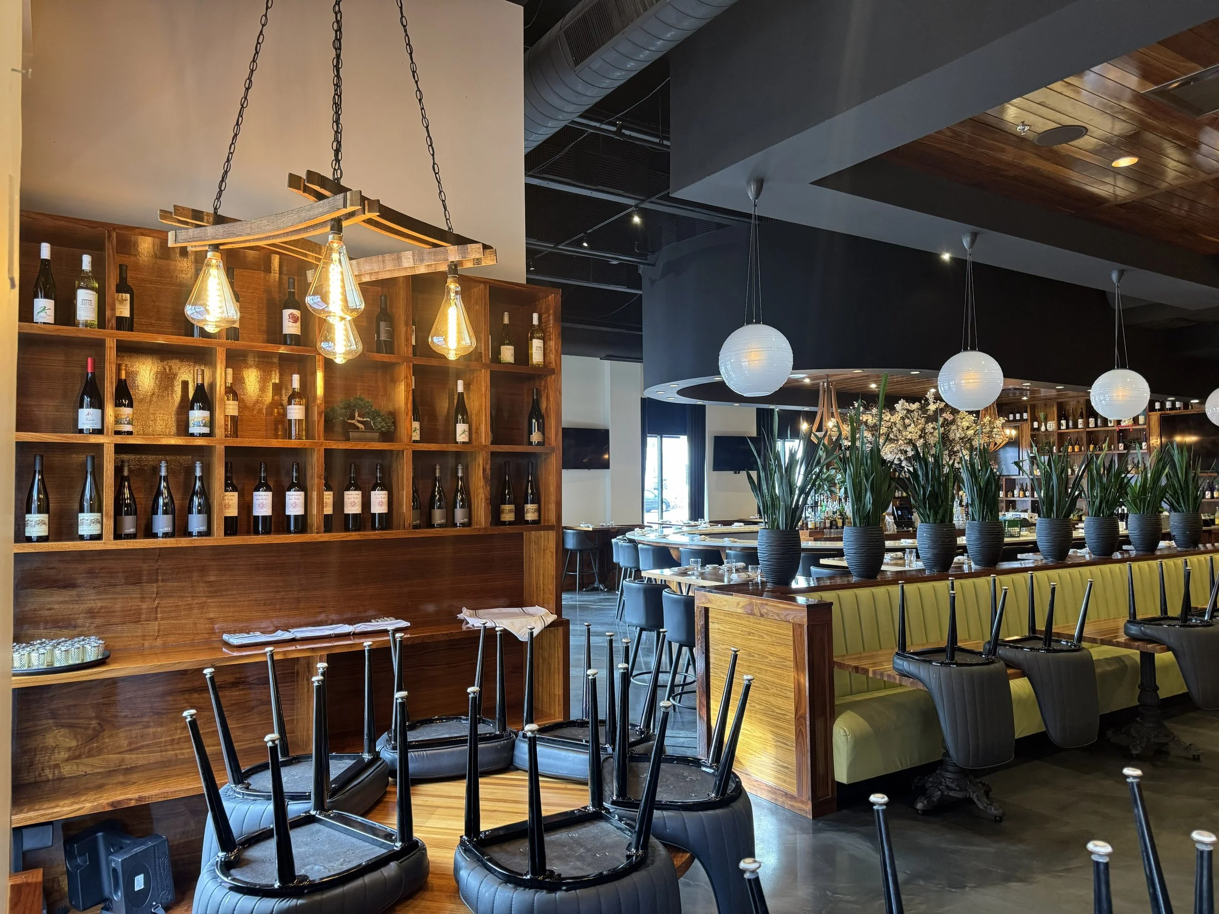 Upside-down chairs stacked on a table in a modern restaurant with wine bottles on wooden shelves, white spherical hanging lights, and potted plants.