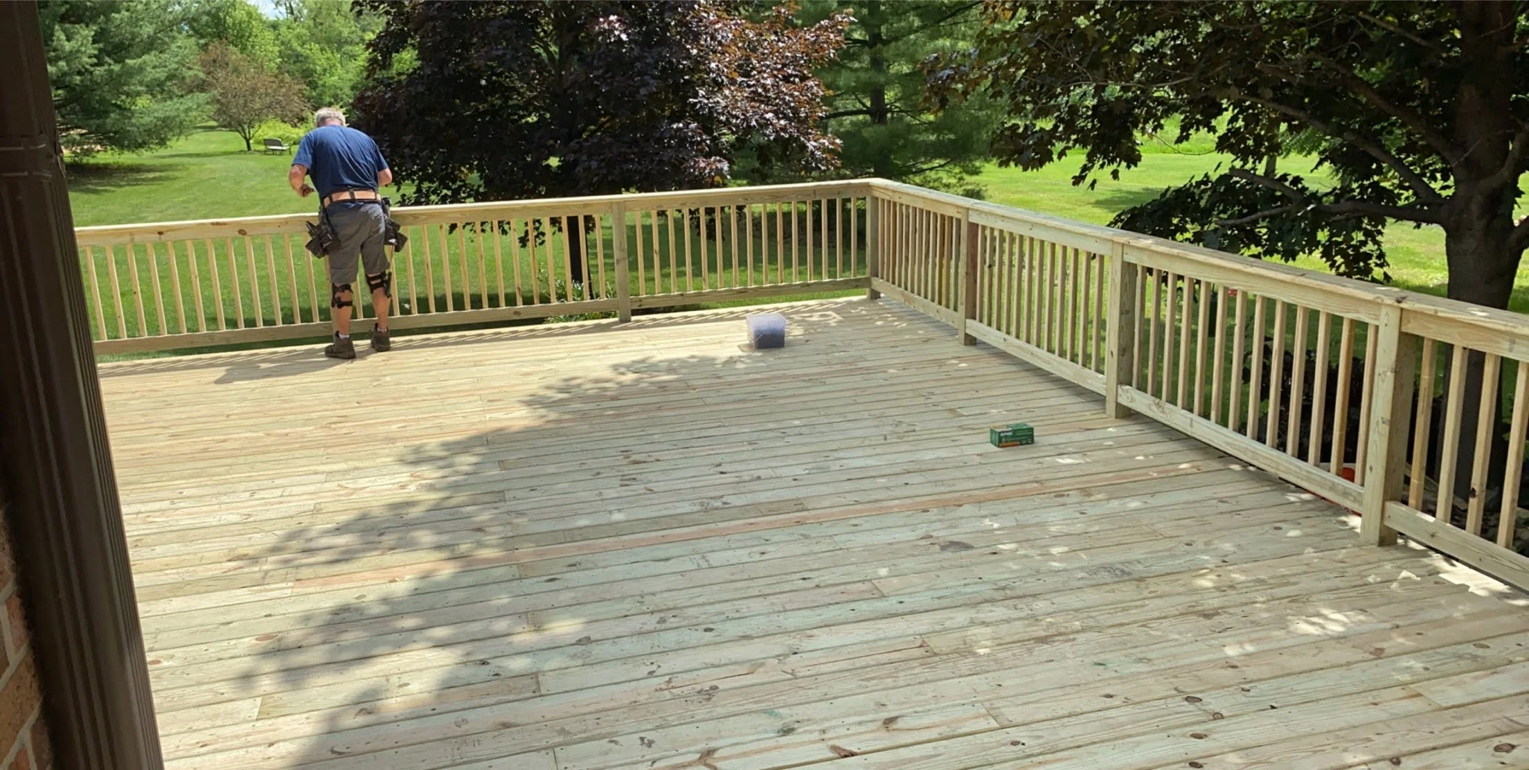 Person working on a wooden deck railing outdoors surrounded by trees and grass on a sunny day. Treated lumber wooden deck.