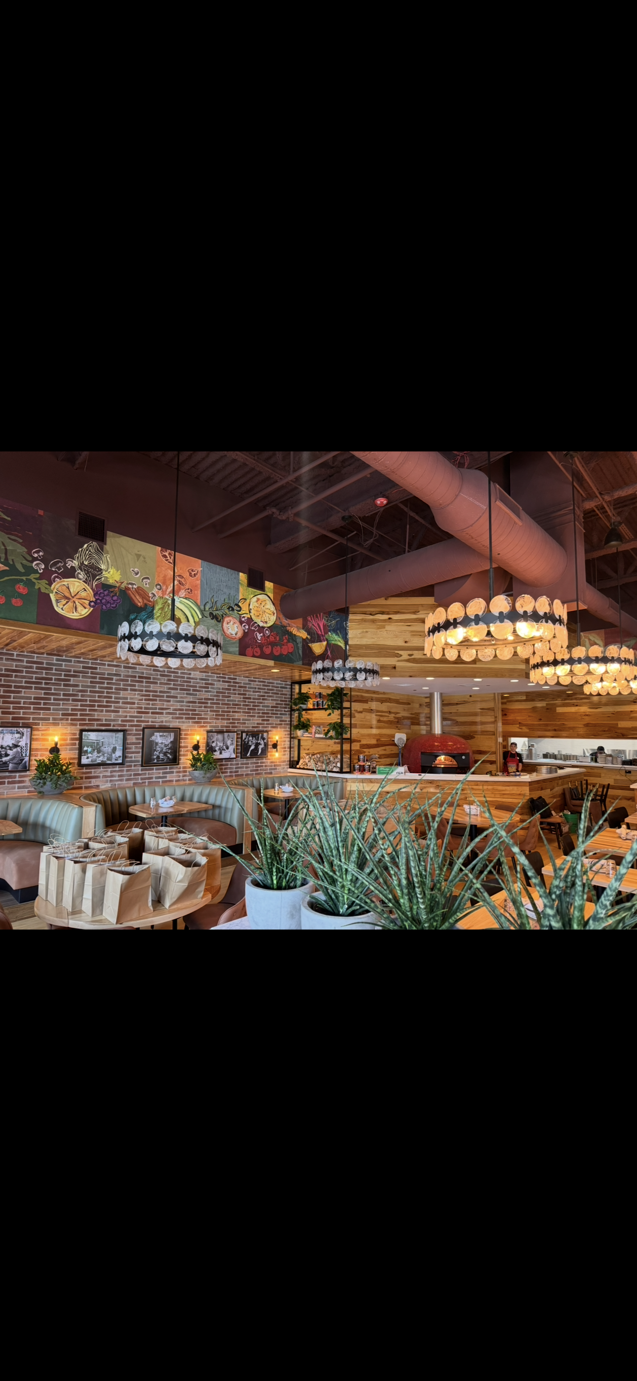 Interior of a modern restaurant with brick and wood walls, potted plants, hanging circular light fixtures, and a large colorful fruit-themed mural.