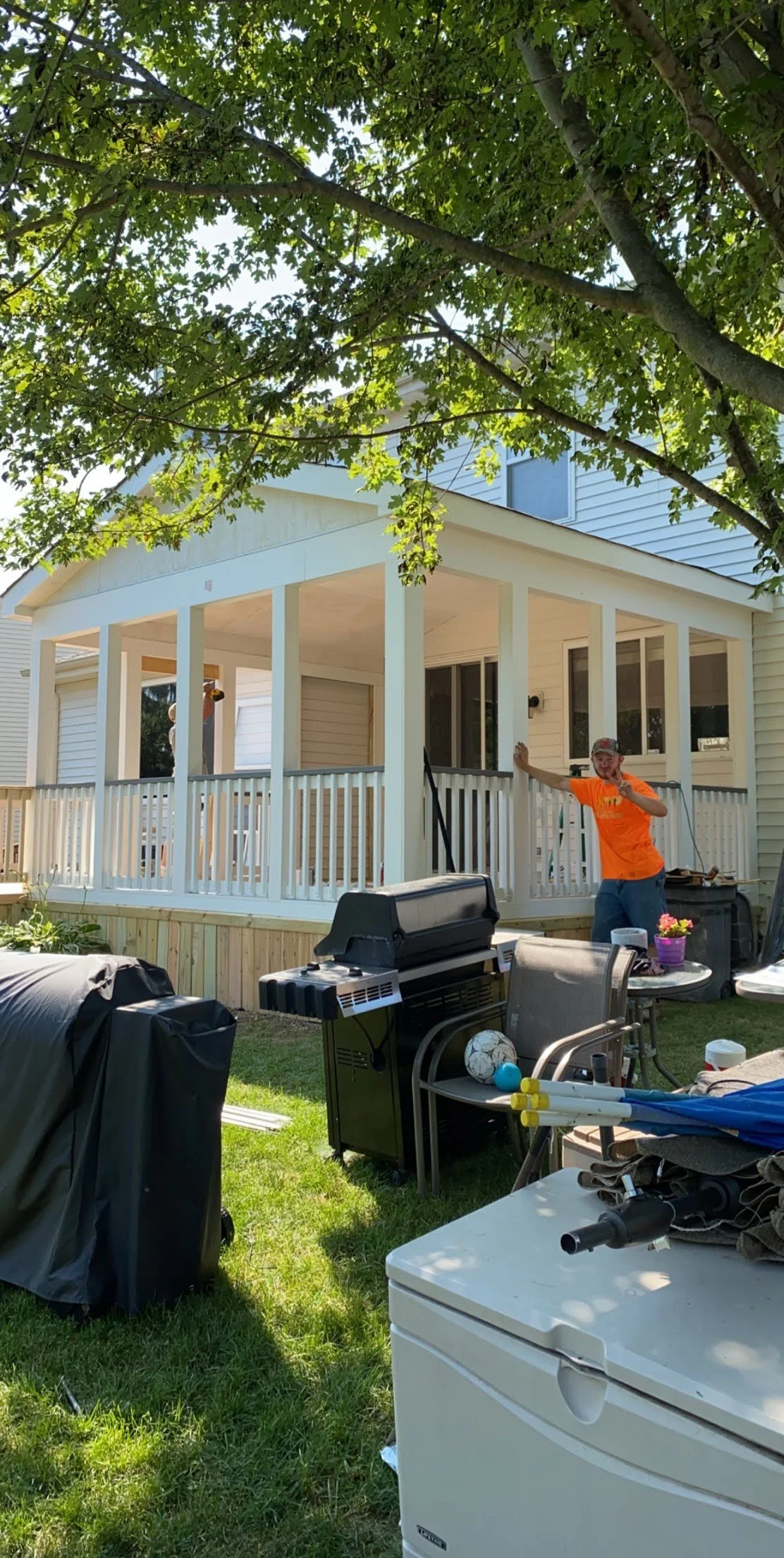 Backyard patio deck near a grill, with outdoor furniture, with a screened porch in the background, shaded by a large tree.