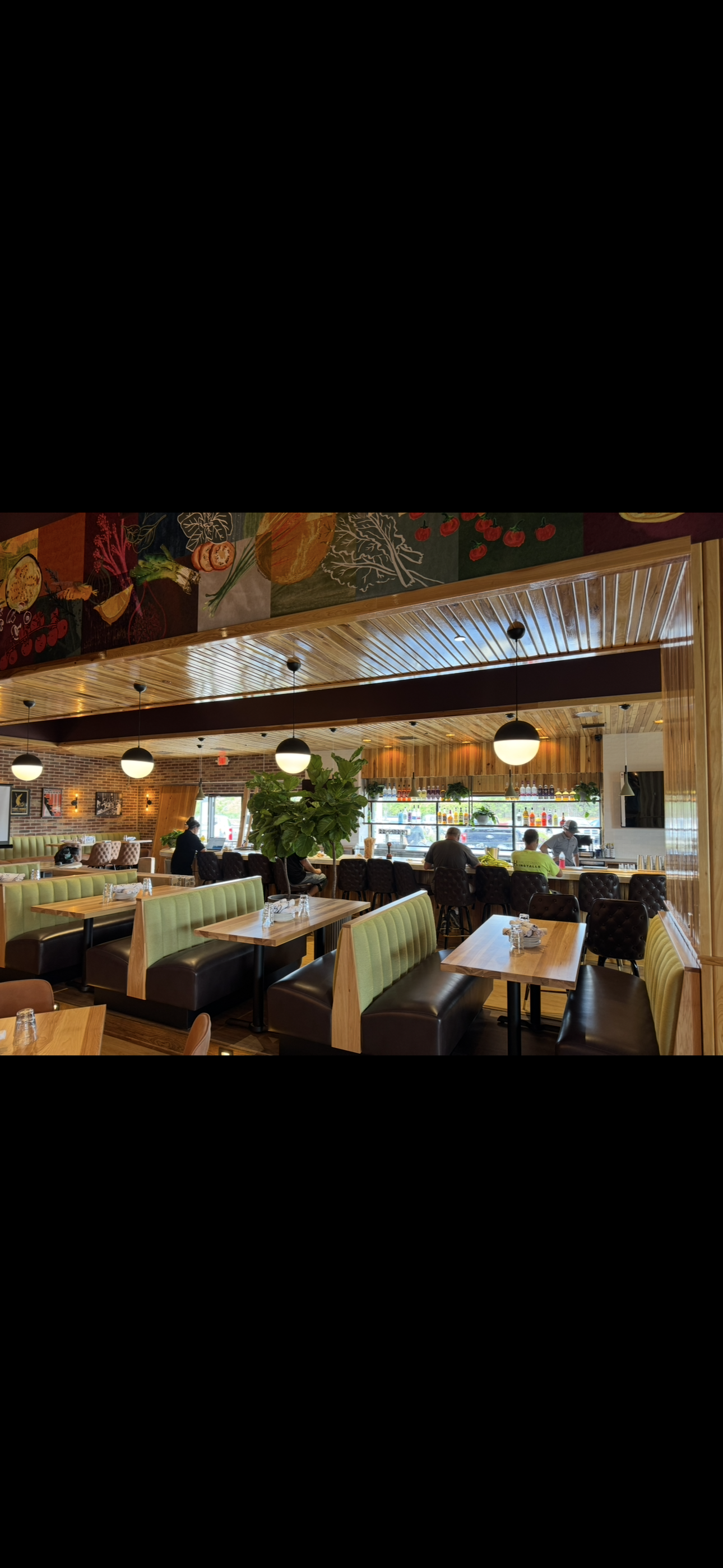 Interior of a restaurant with wooden ceiling, hanging pendant lights, a bar area with bottles and glasses, green booth seating, tables with napkins and utensils, a large green plant, and customers seated at the bar and tables.