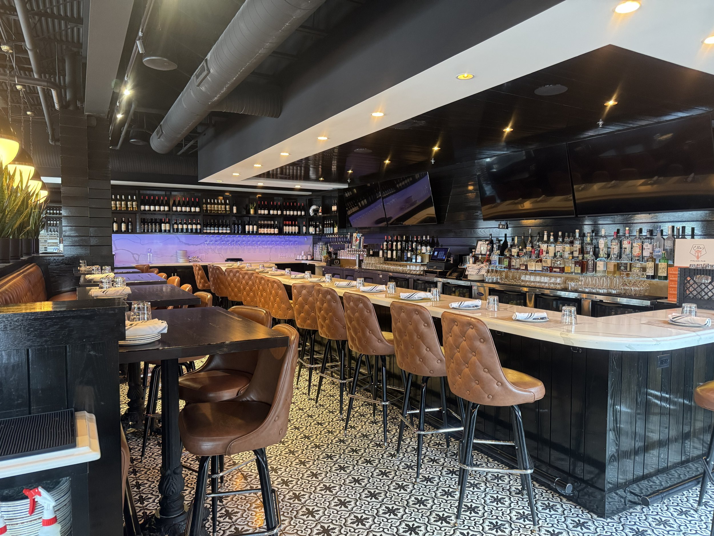 Interior of a restaurant or bar with a bar counter, several bar stools, a wine wall, and television screens on a dark wood wall.