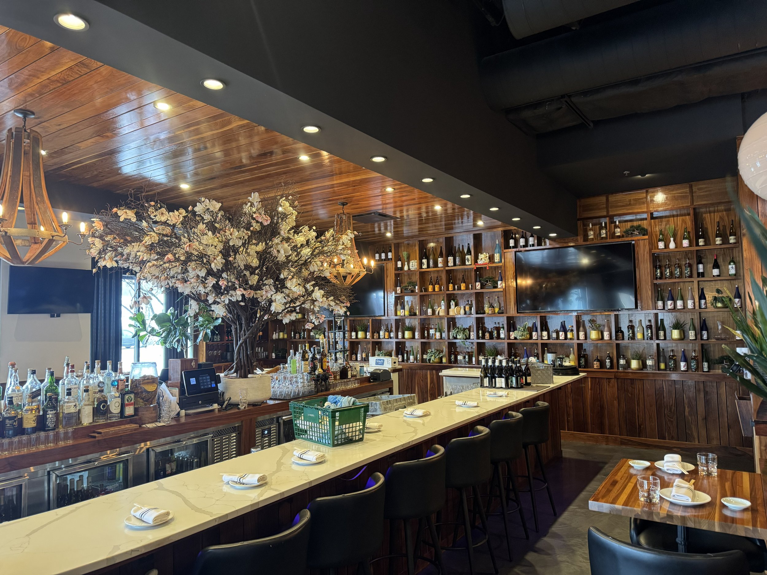 Interior of a bar with a large floral centerpiece, wooden shelves filled with bottles, a flat screen TV, a marble bar counter with bar stools, and dining tables prepared with plates, napkins, and glasses.