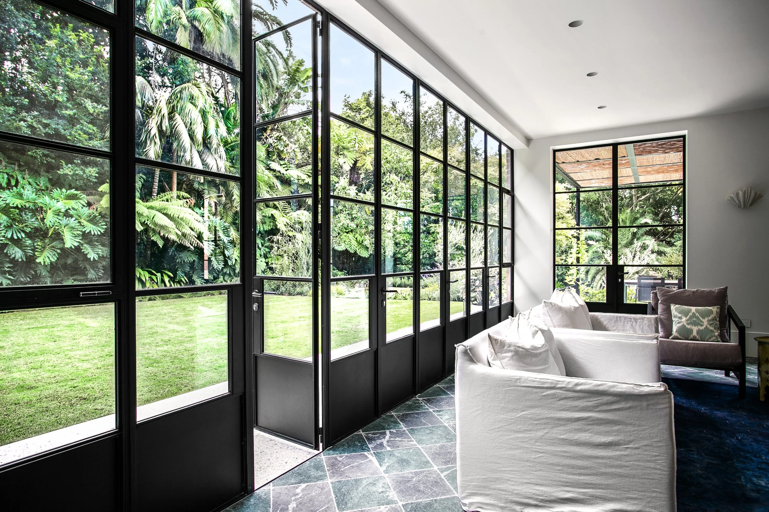 Interior view of a bright living room with large black-framed glass doors and windows opening to a lush green garden with trees.