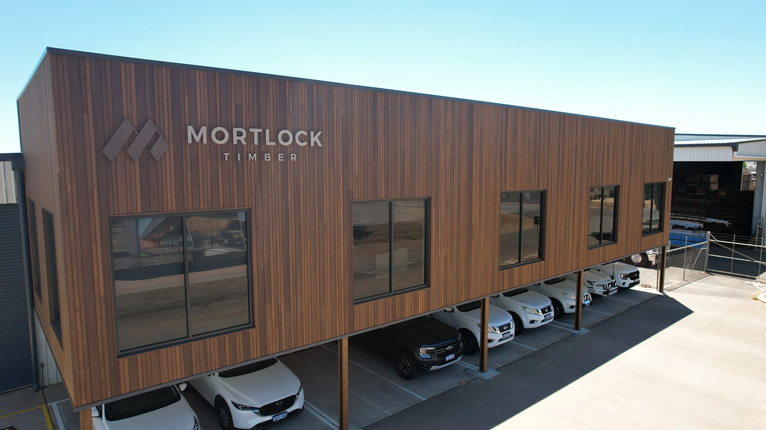 A two-story commercial building with a wooden exterior and large windows, labeled 'Mortlock Timber,' with cars parked underneath on a sunny day.