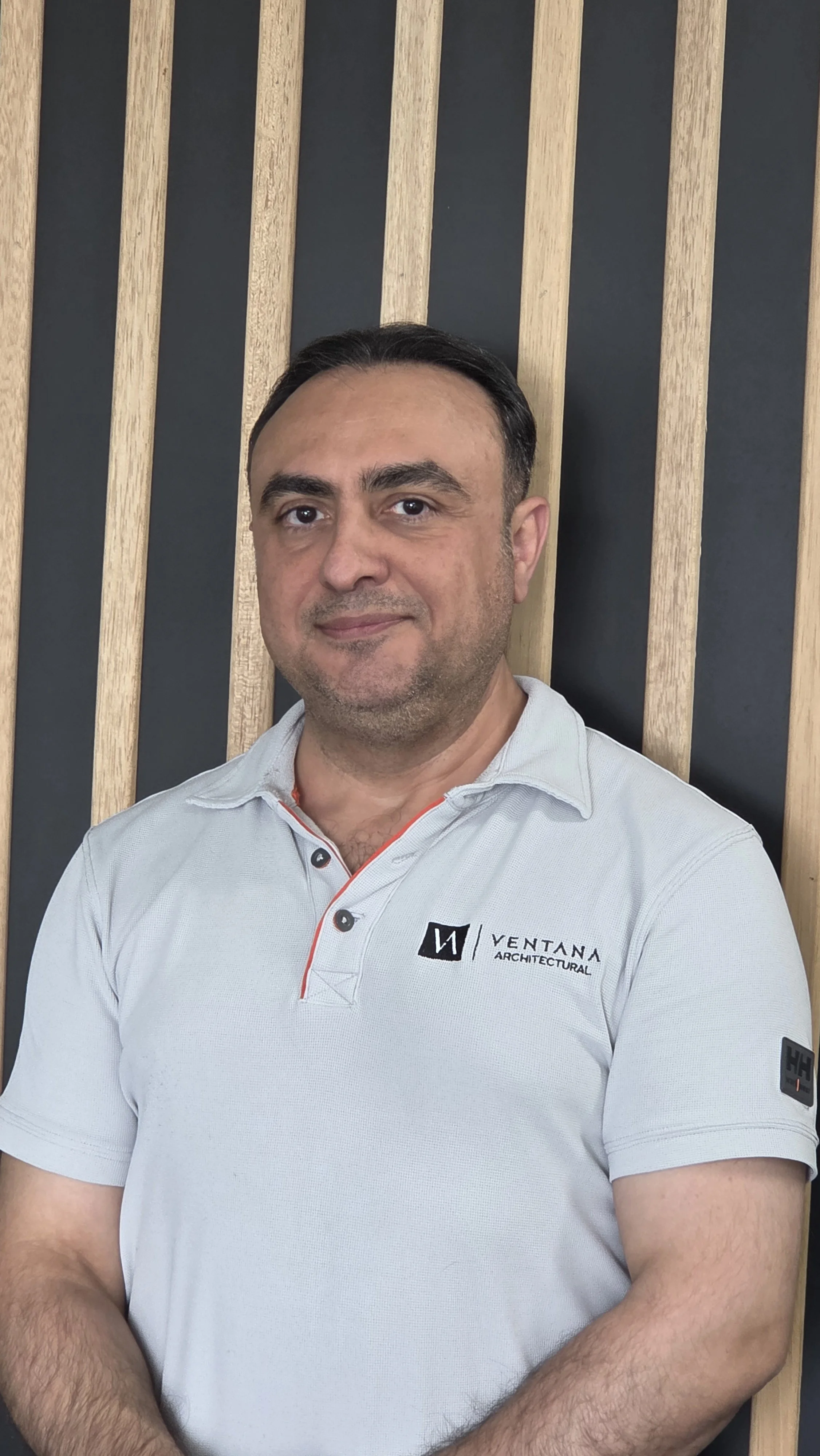 A man with dark hair and a short beard wearing a white polo shirt with the logo and text "Ventana Architectural" standing against a wall with vertical wooden slats.