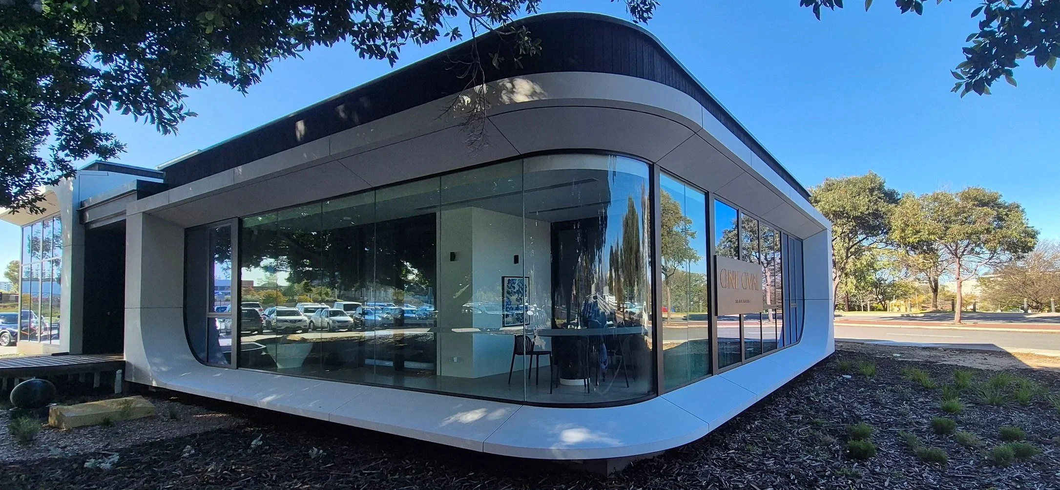 Modern building with curved glass facade, situated in a landscaped area with trees, parked cars reflected in the windows, and a clear blue sky overhead.