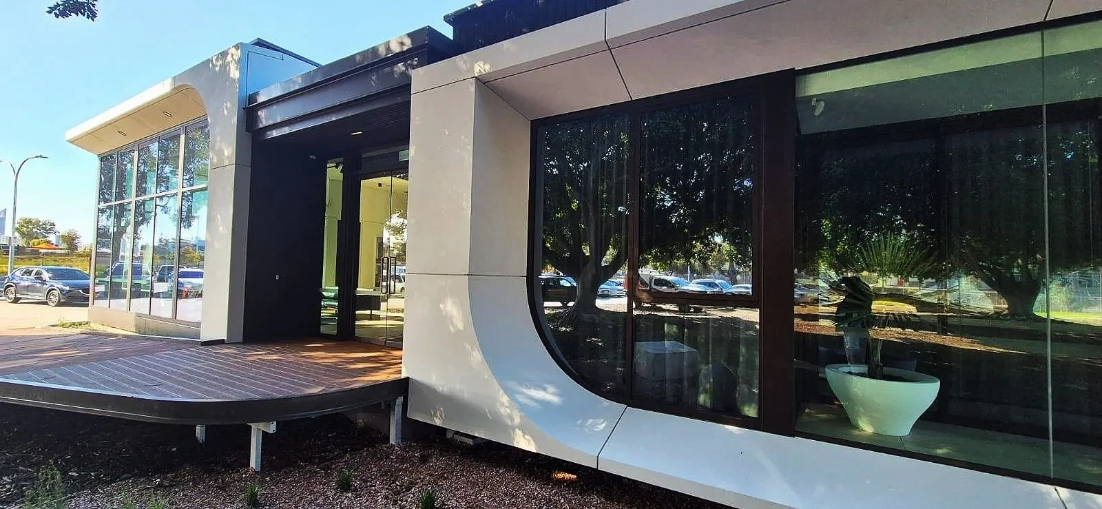 Modern commercial building exterior with large glass windows, automatic sliding glass doors, wooden deck, and trees reflected in the windows
