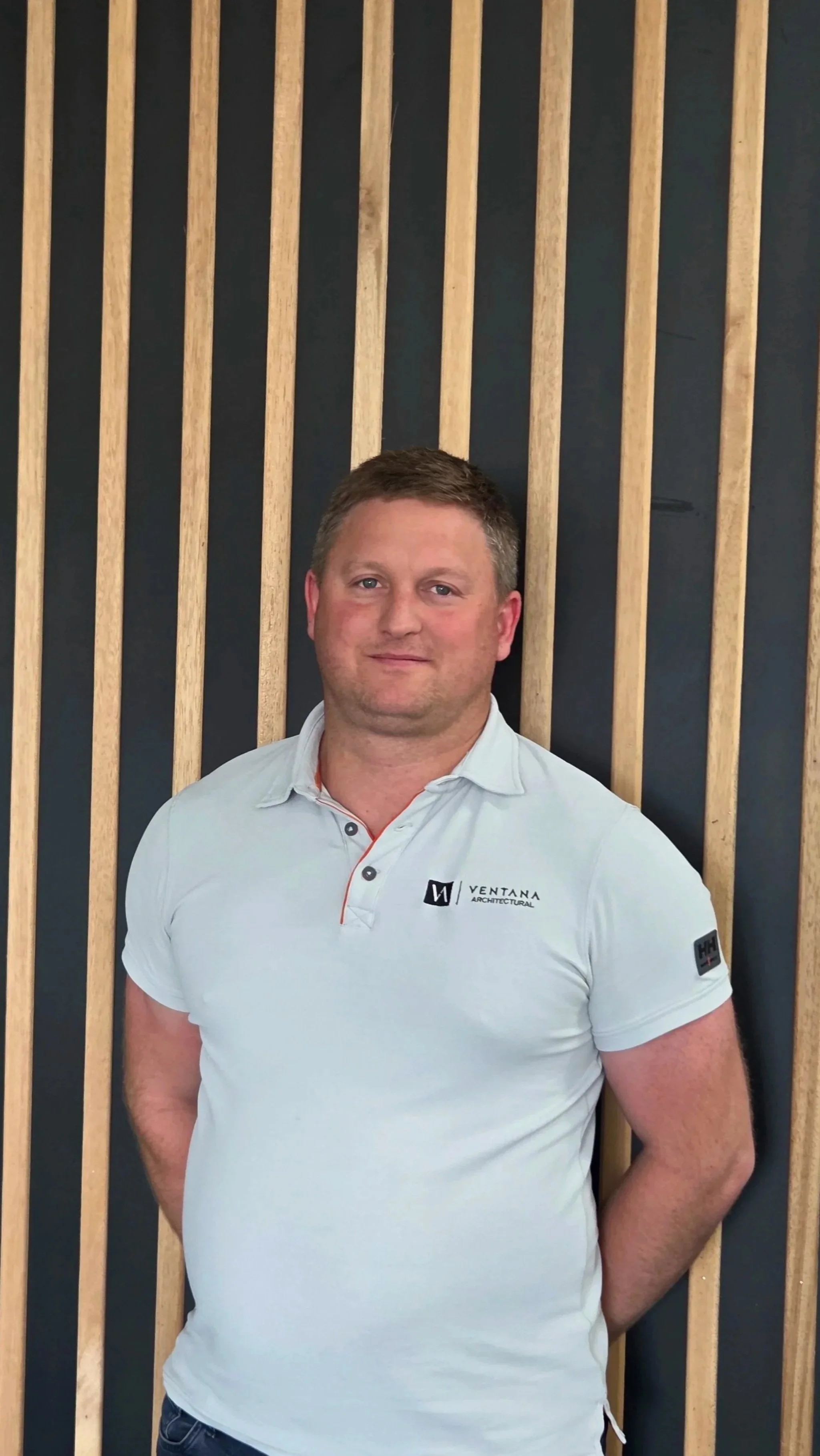 A man standing in front of a wall with vertical wooden slats, wearing a white polo shirt with the words 'Ventana Architecture' on it.