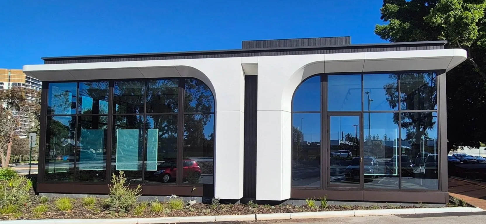 Modern building with large glass windows and white walls, surrounded by a parking lot and greenery, with a clear blue sky in the background.