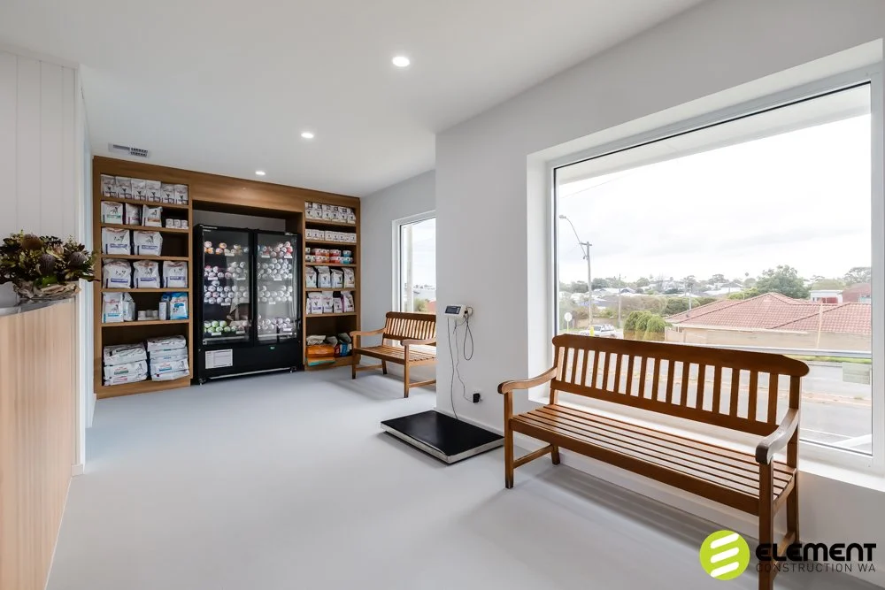 A clean, well-lit waiting area with two wooden benches, a large window with a view of houses and trees, and a storage unit with linens and towels. A computer scale is on the floor near the window.