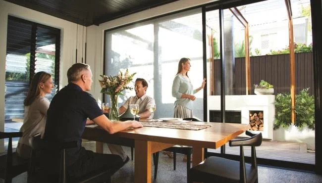 A family of four sitting around a dining table inside a modern house, with a woman standing near glass doors opening to a patio with firewood and plants.