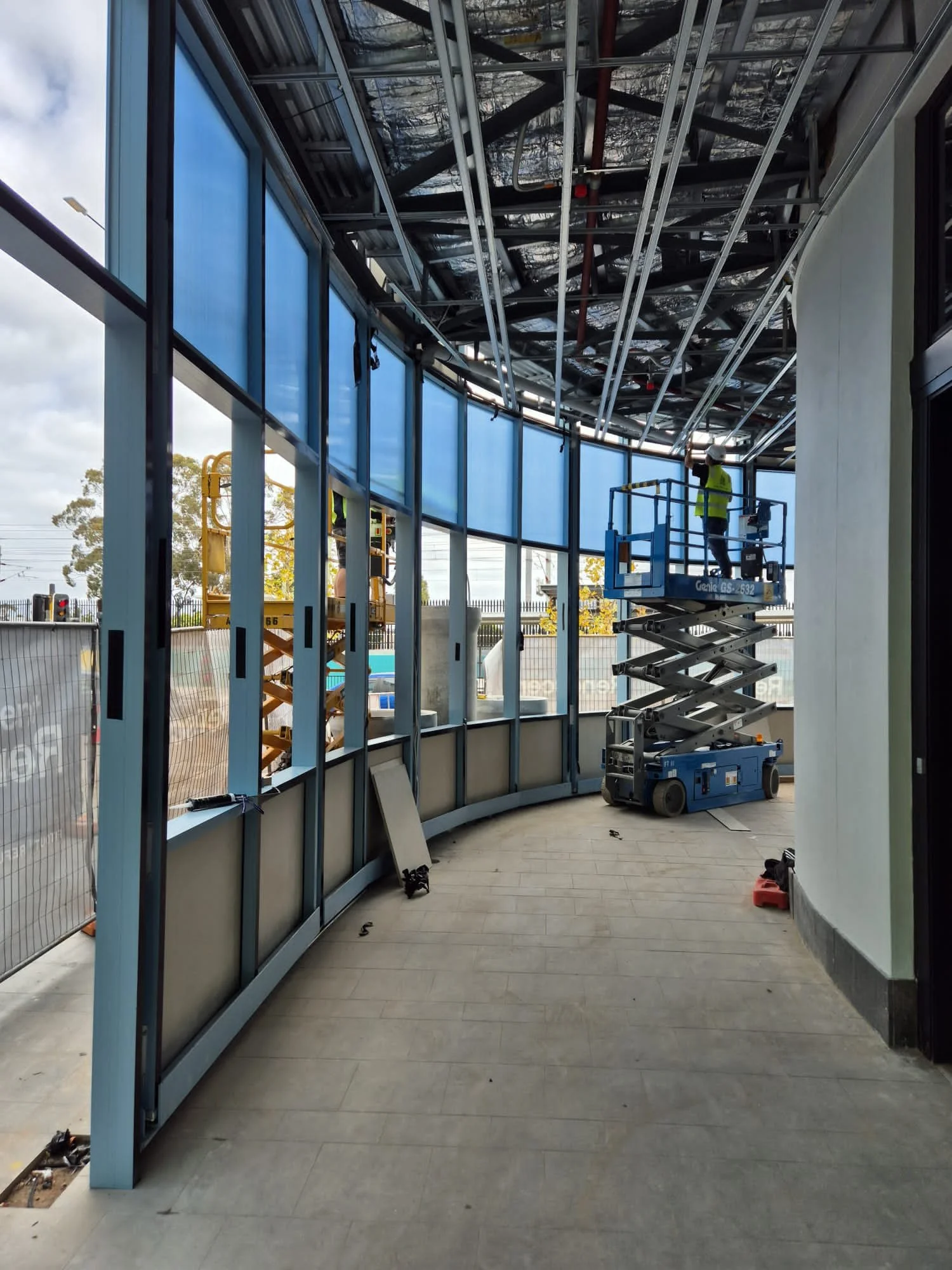 Construction workers in a lift working on a curved glass exterior wall of a building, with construction materials and equipment around.