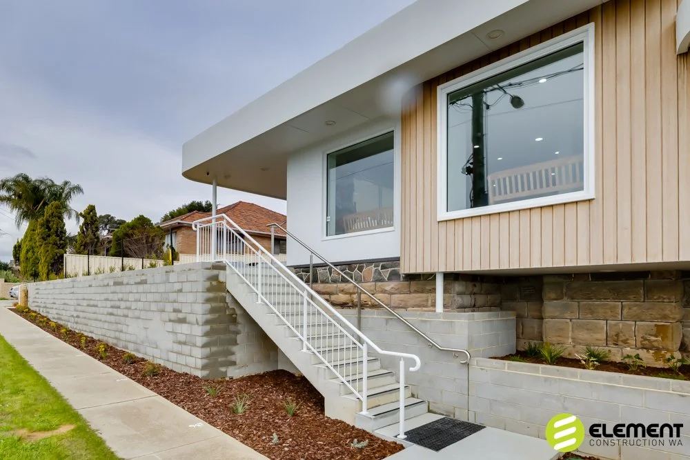 Modern house with front steps and white railing, brick and wood exterior, large windows, landscaped yard, and sidewalk.