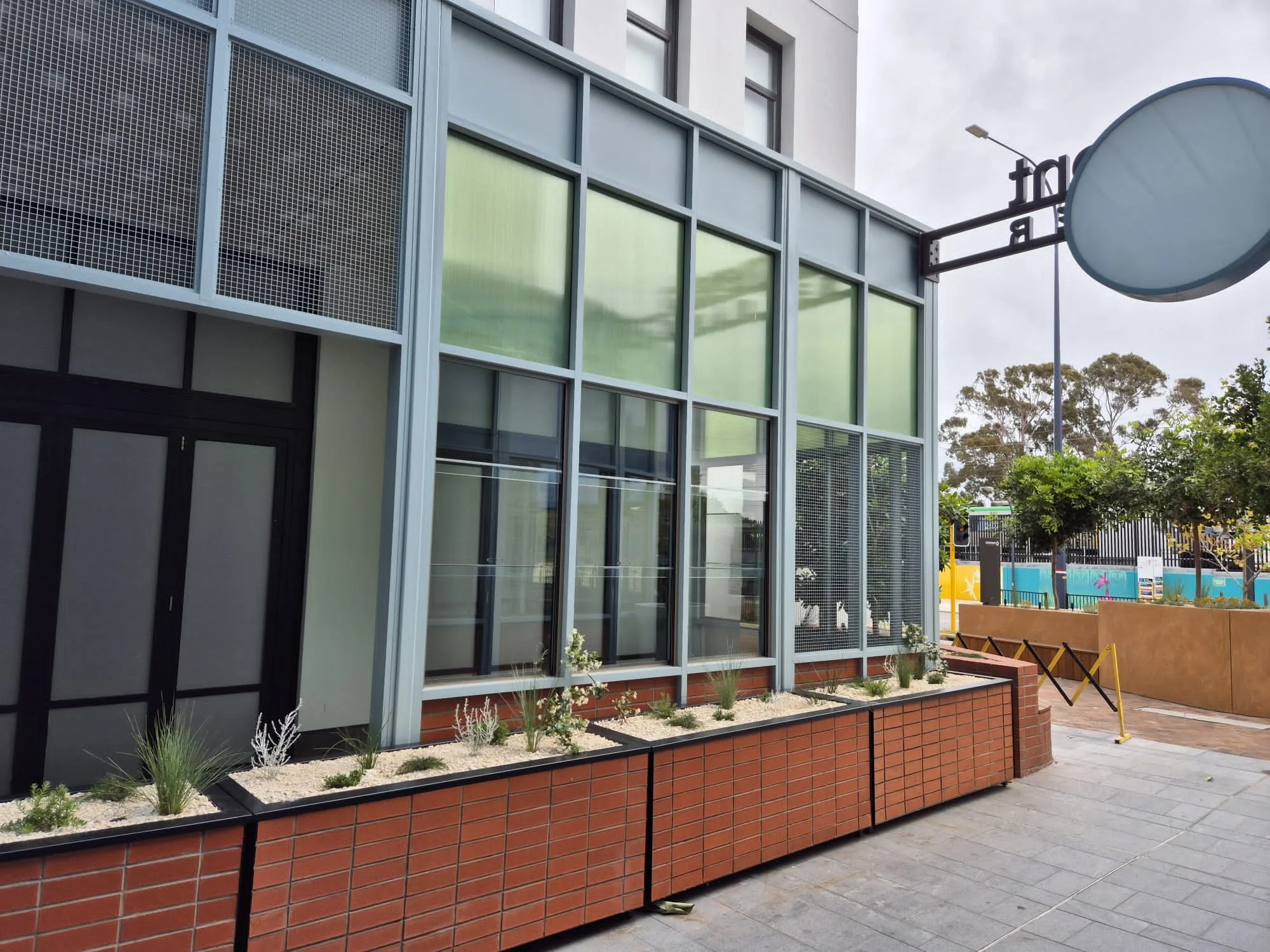Exterior view of a modern building with large glass windows, brick planters with small plants in front, and a sidewalk. Part of a round sign is visible on the right, and trees and a fence are in the background.