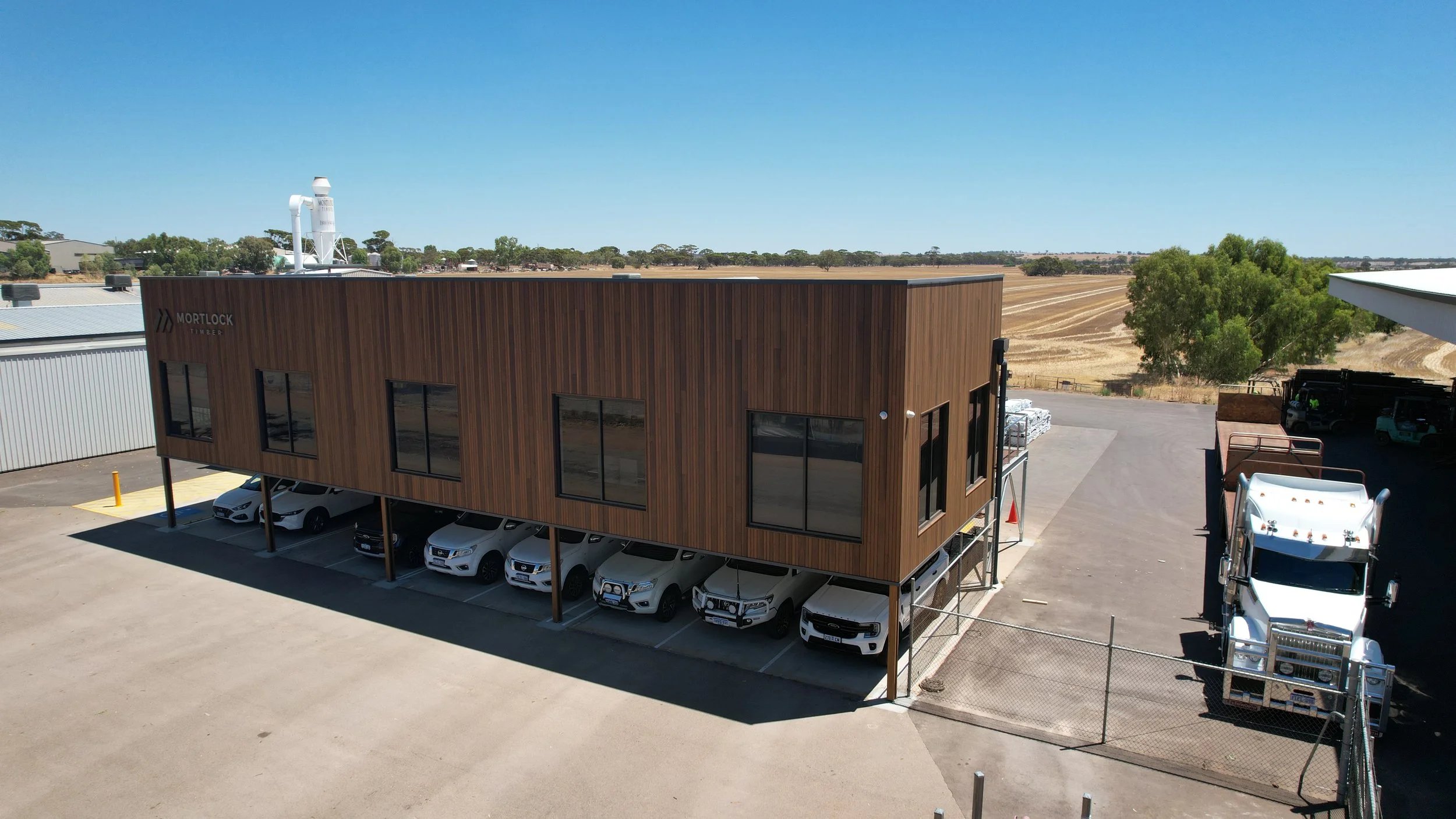 A two-story commercial building with a wood exterior, parked cars underneath, and a large white truck beside it. The building has the name 'Mortlock Timber' on the upper left corner and is situated in an industrial area with farmland in the backgroun
