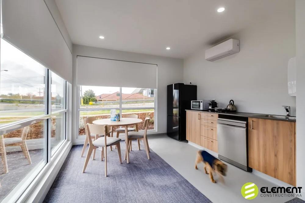 Modern kitchen and dining area with large windows, round dining table with four chairs, black refrigerator, microwave, toaster, electric kettle, dishwasher, wooden cabinets, and a small dog walking inside.