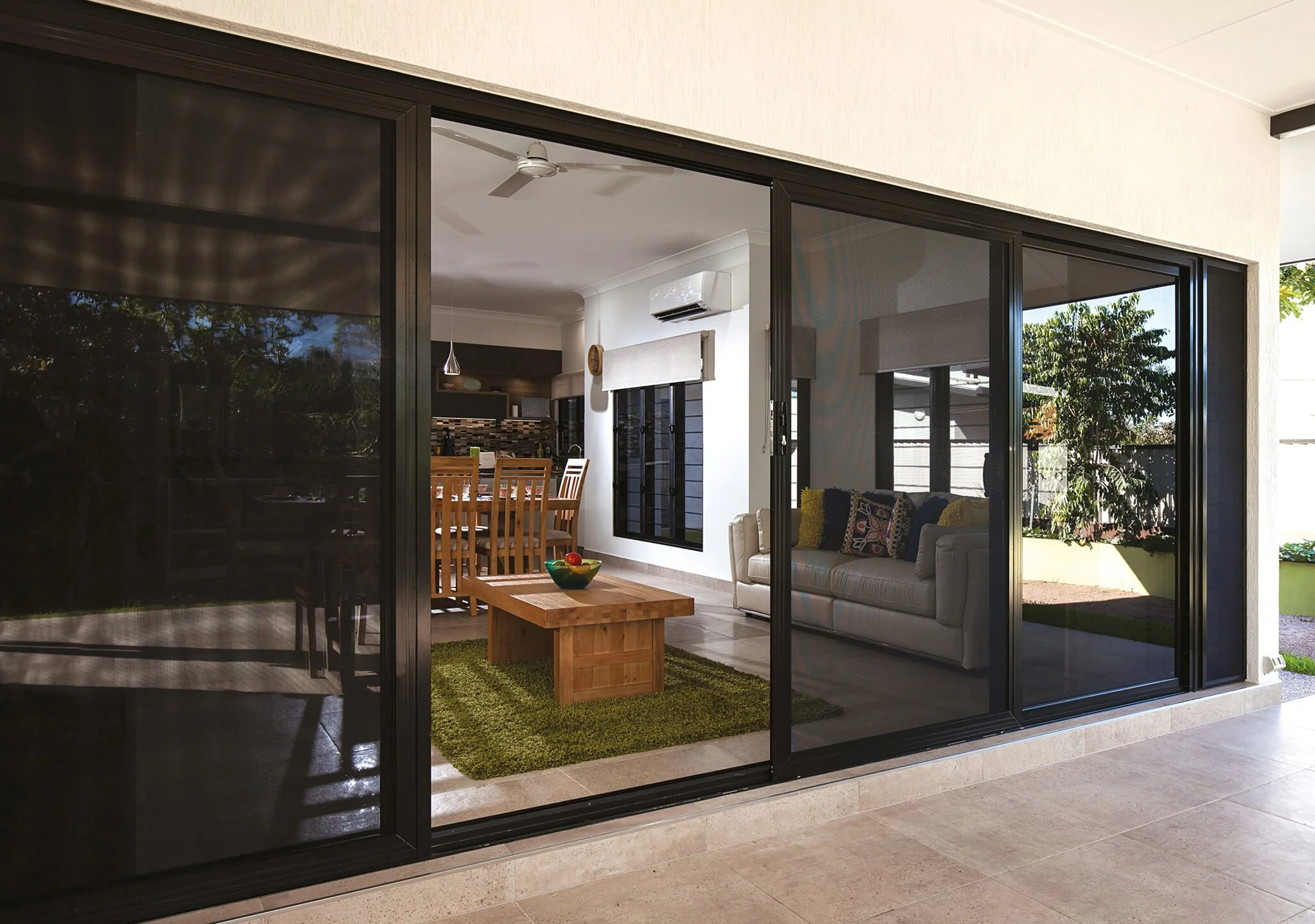 Sliding glass doors leading to a living room with a beige sofa, wooden dining table with chairs, small coffee table, and a green rug, with a backyard visible outside.