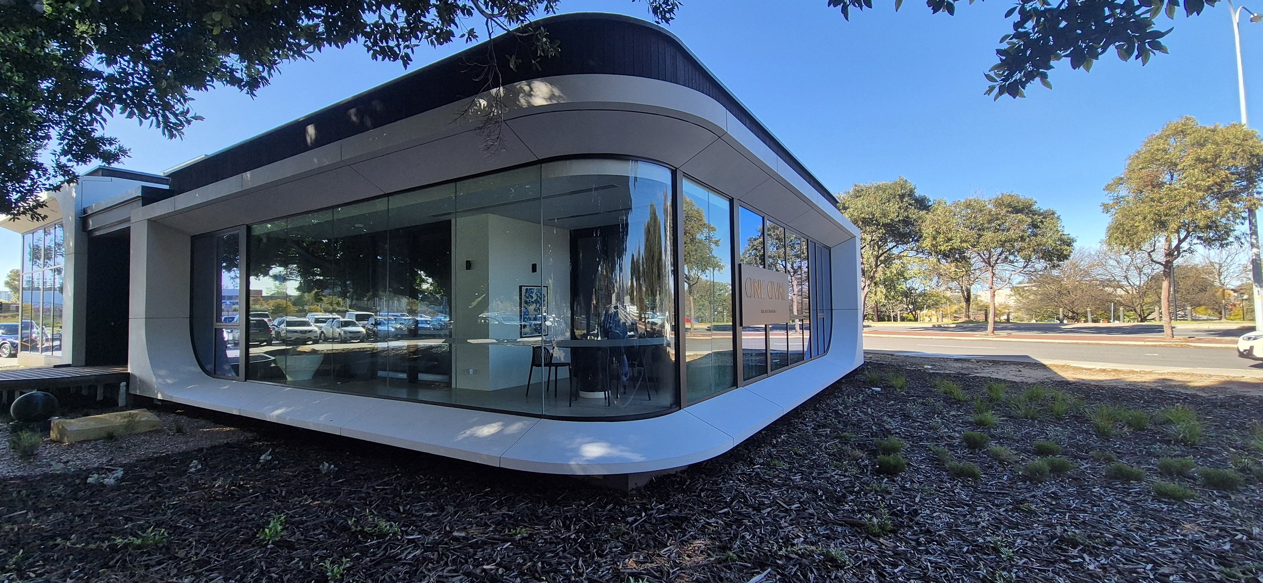 Modern glass building with curved design and trees in the background, parked cars visible through the glass, and a sign inside that says 'Only One'.