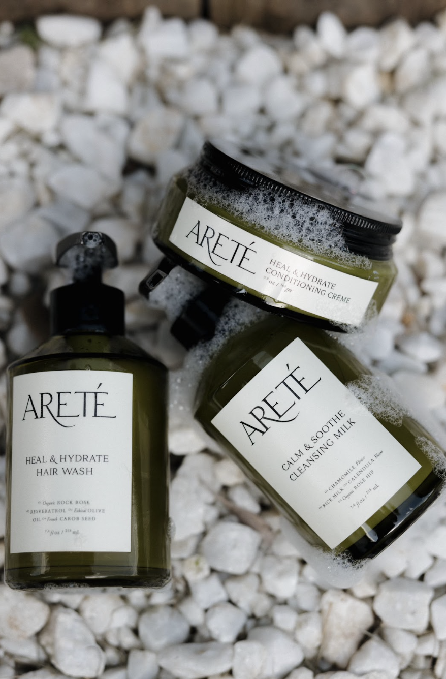Three green bottled skincare products on white rocks: Areté Heal & Hydrate Hair Wash, Areté Heal & Hydrate Conditioning Crème, and Areté Calm & Soothe Cleansing Milk.