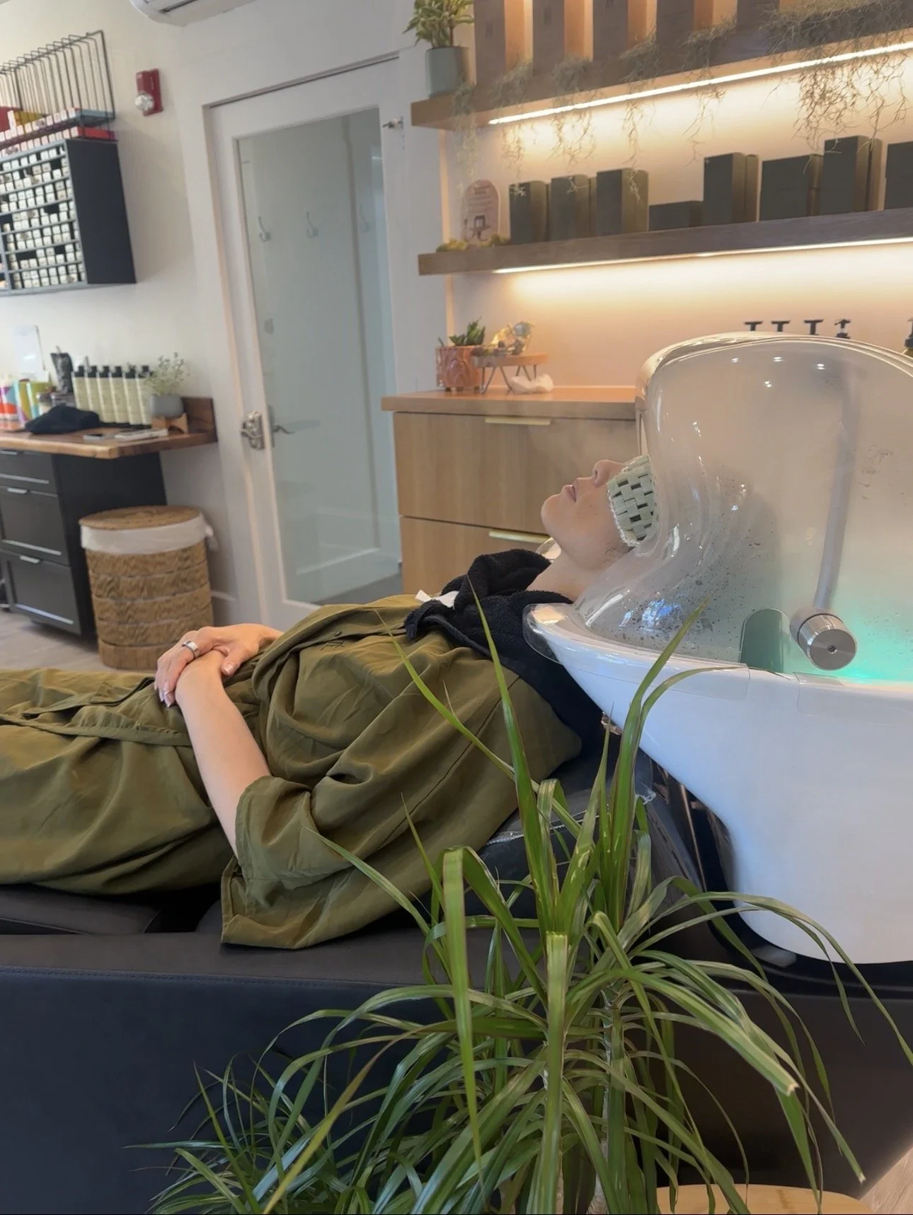 A woman lying on a treatment bed with her eyes closed in a spa or wellness center, with a neck and shoulder massage device around her neck, surrounded by indoor plants and modern decor.