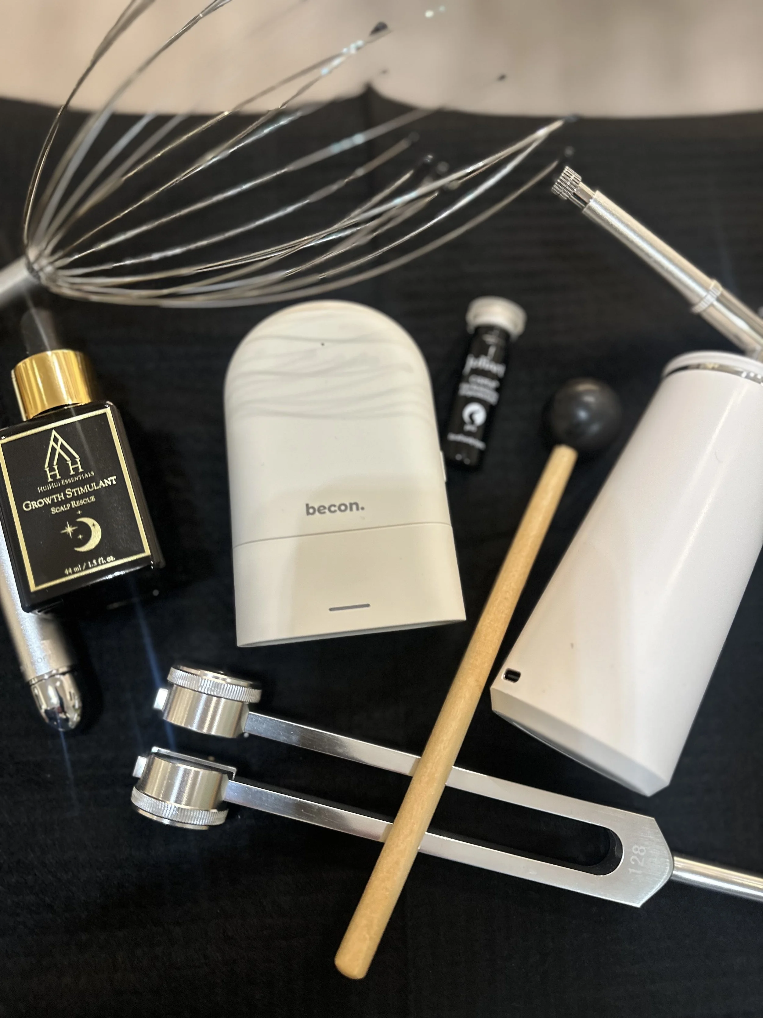 Collection of beauty and skincare items including perfume, white and black bottles, a small tube, a facial device, a wooden applicator, and a metal tool with adjustable dials, arranged on a black surface.