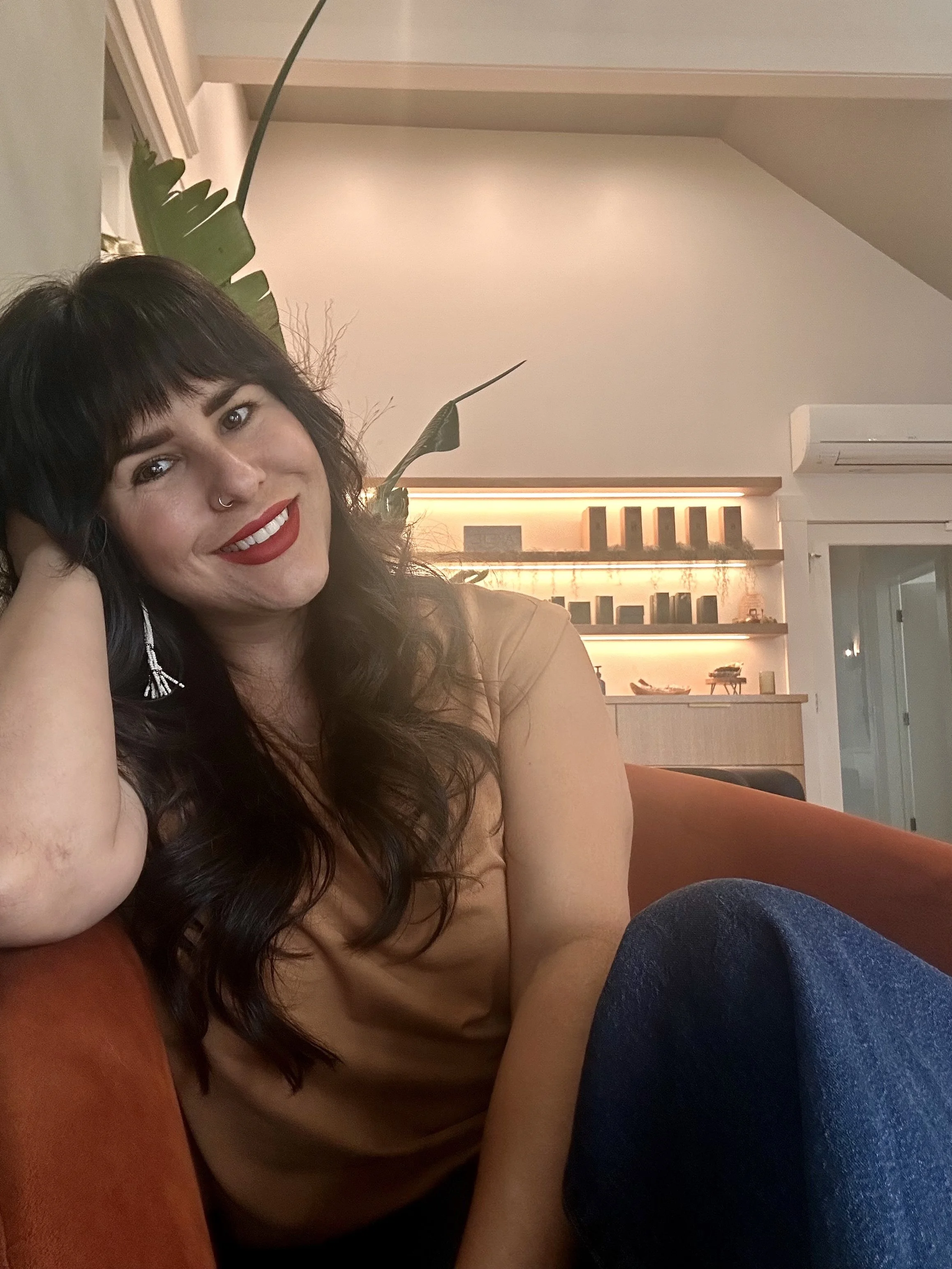 A woman with black hair, red lipstick, and a nose ring lounging on a red couch in a modern living room with wooden shelves, plants, and warm lighting.
