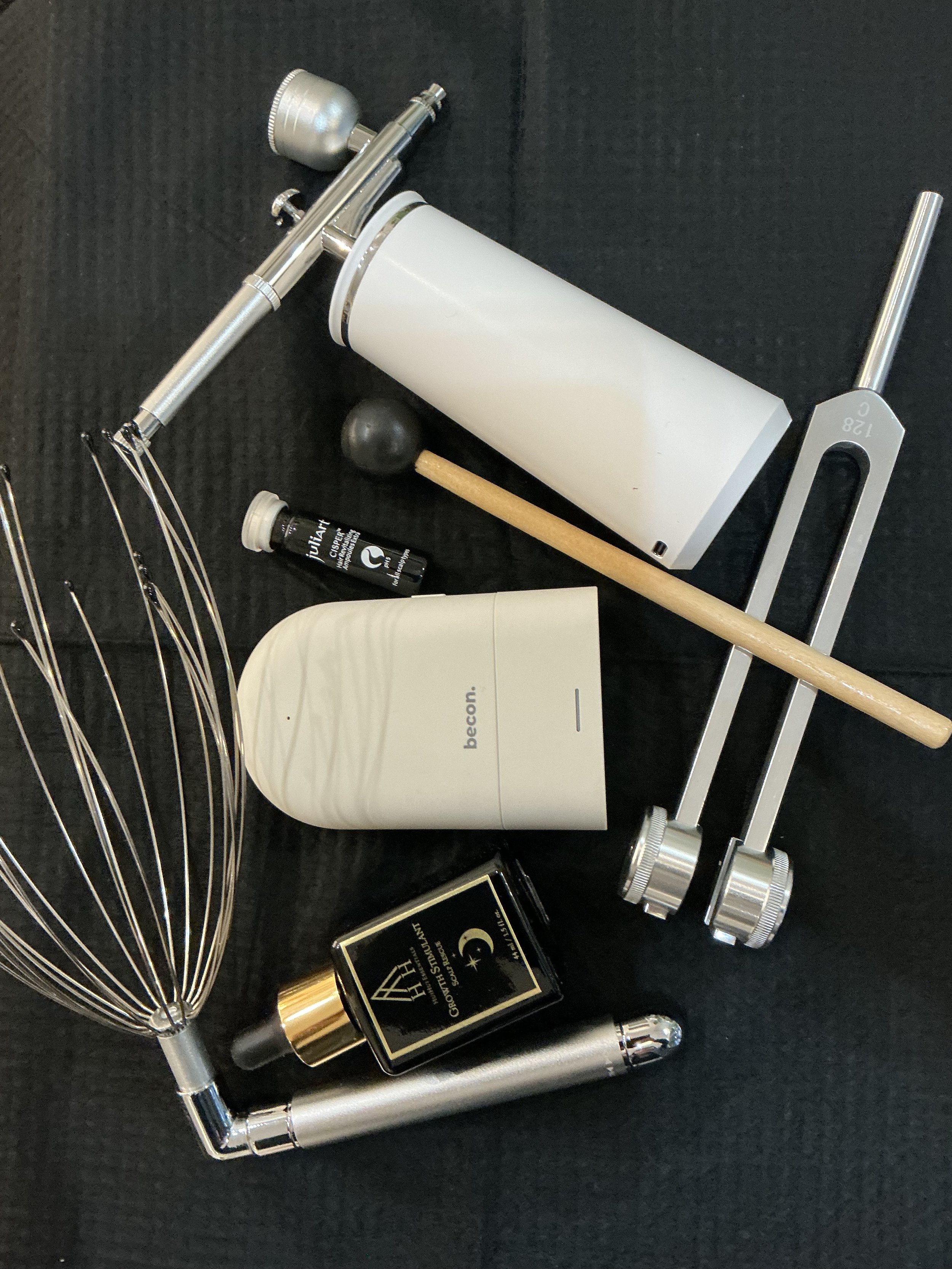 Beauty tools including a stainless steel hairbrush, a white tube of skincare product, a small bottle of 'Glow' serum, a black box of 'Growth Serum' with gold accents, a black and gold roller, and a silver metal tool with measurements, arranged on a black surface.