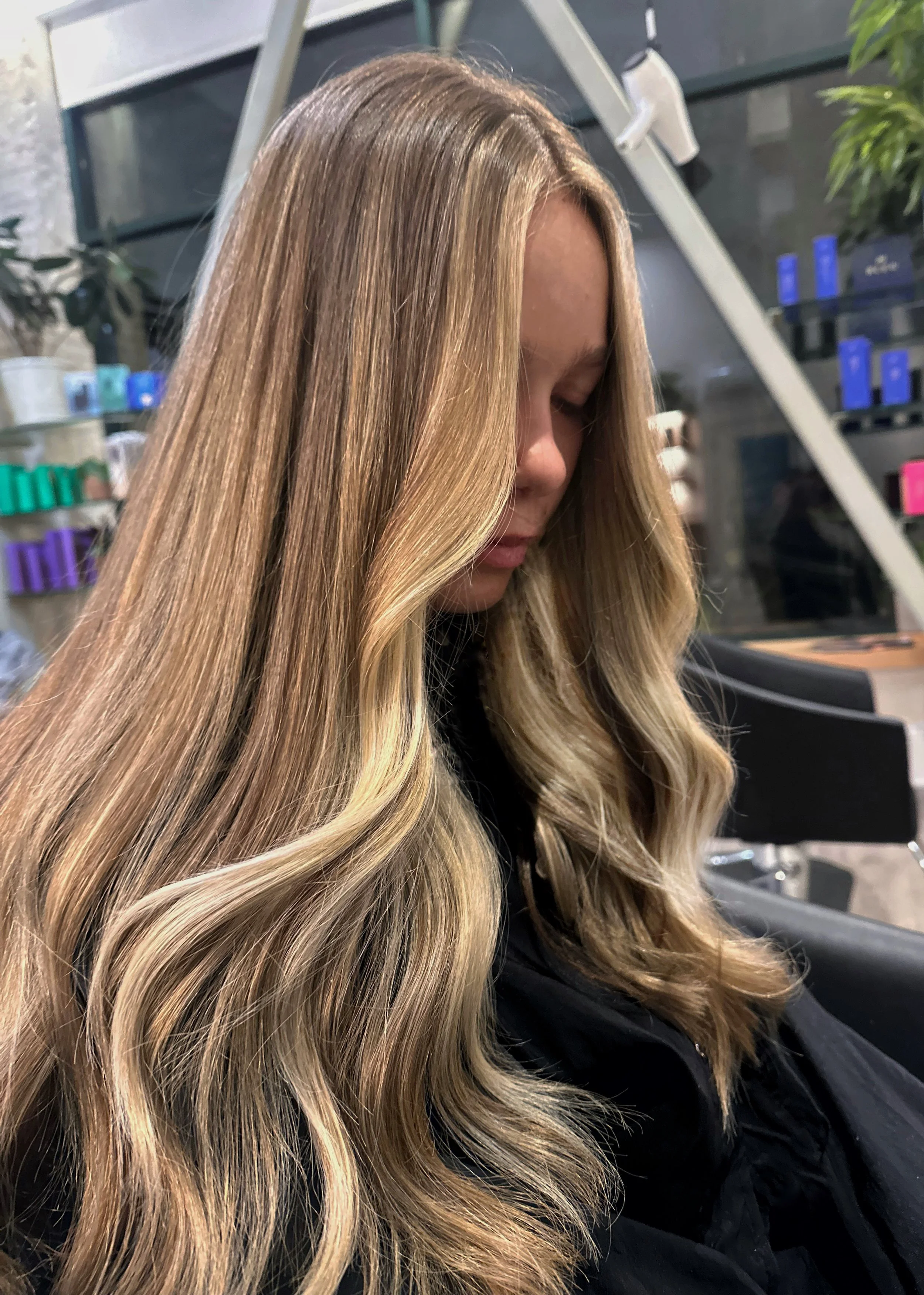 Woman with long blonde hair sitting in a salon chair, with shelves of hair care products in the background.