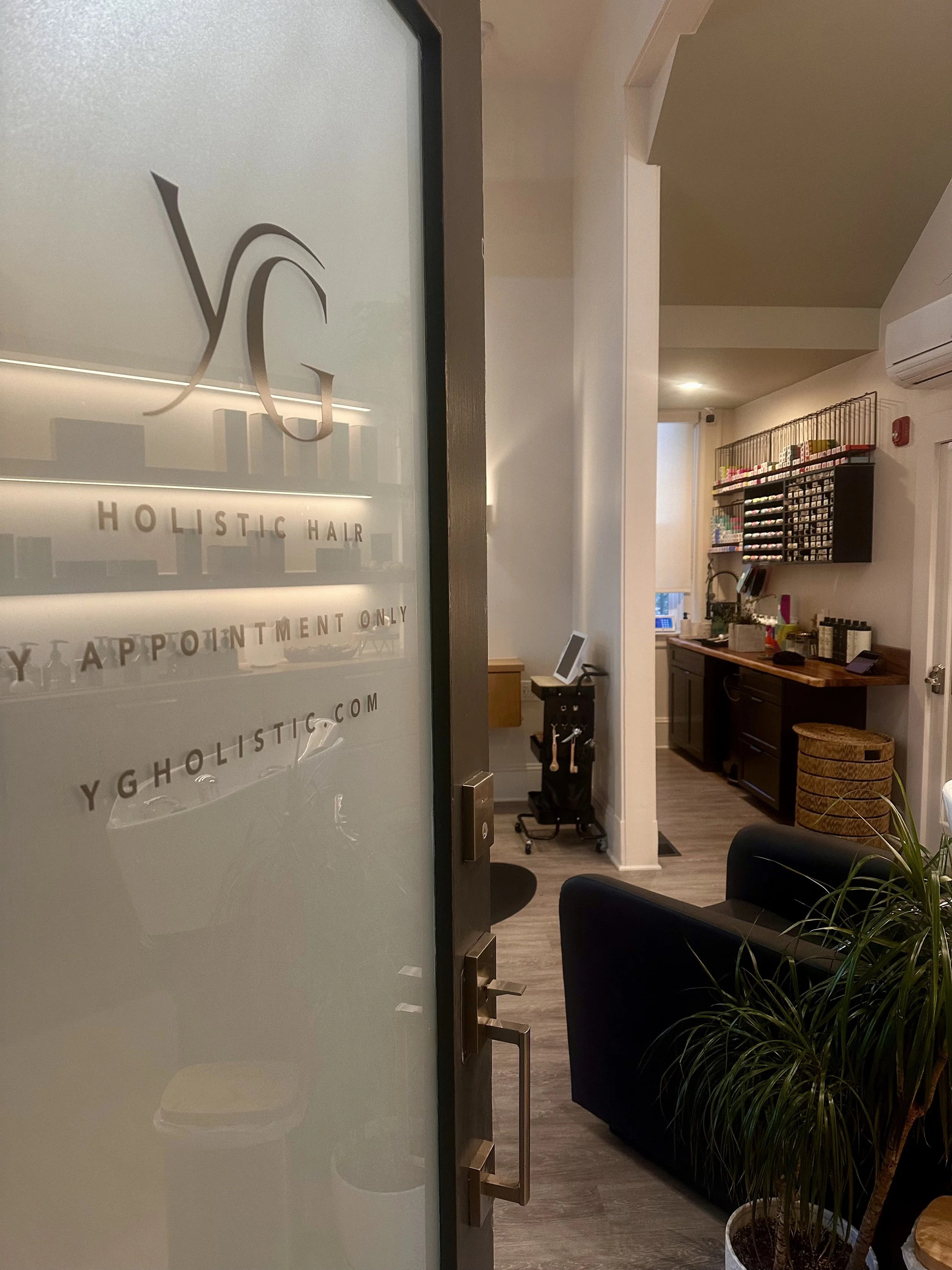 Interior view of a hair salon with a reception desk, wall-mounted storage of hair products, and a waiting area with black chairs and a potted plant.