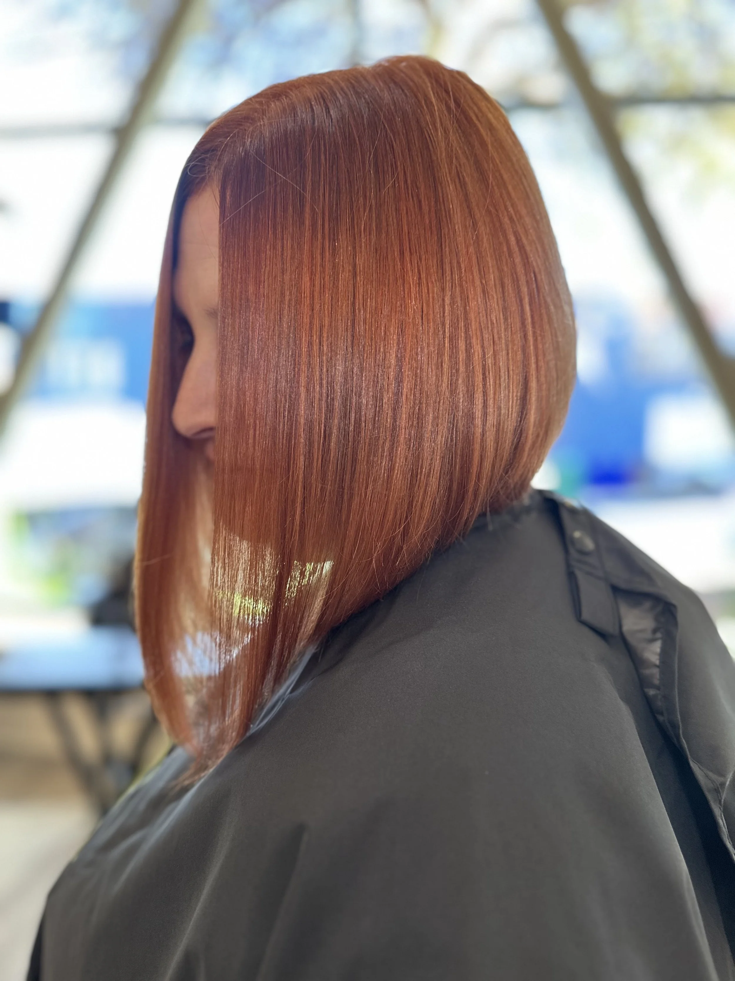 Close-up of a woman with straight, shiny, red hair, wearing a black outfit, indoors with a blurred background.
