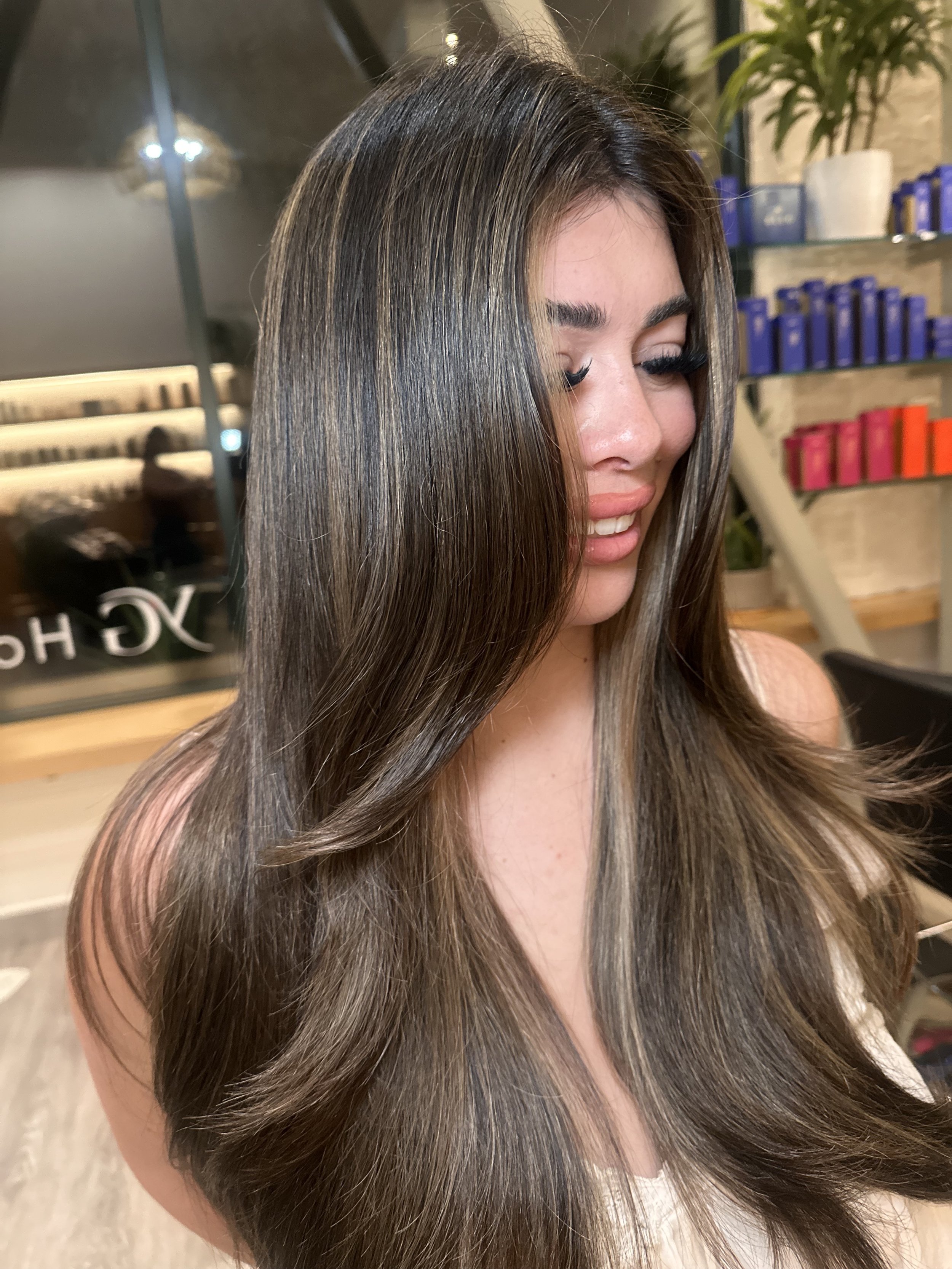 Close-up of a woman with long, shiny, brown hair with blonde highlights, smiling and looking down, in a salon setting with hair products on shelves in the background.