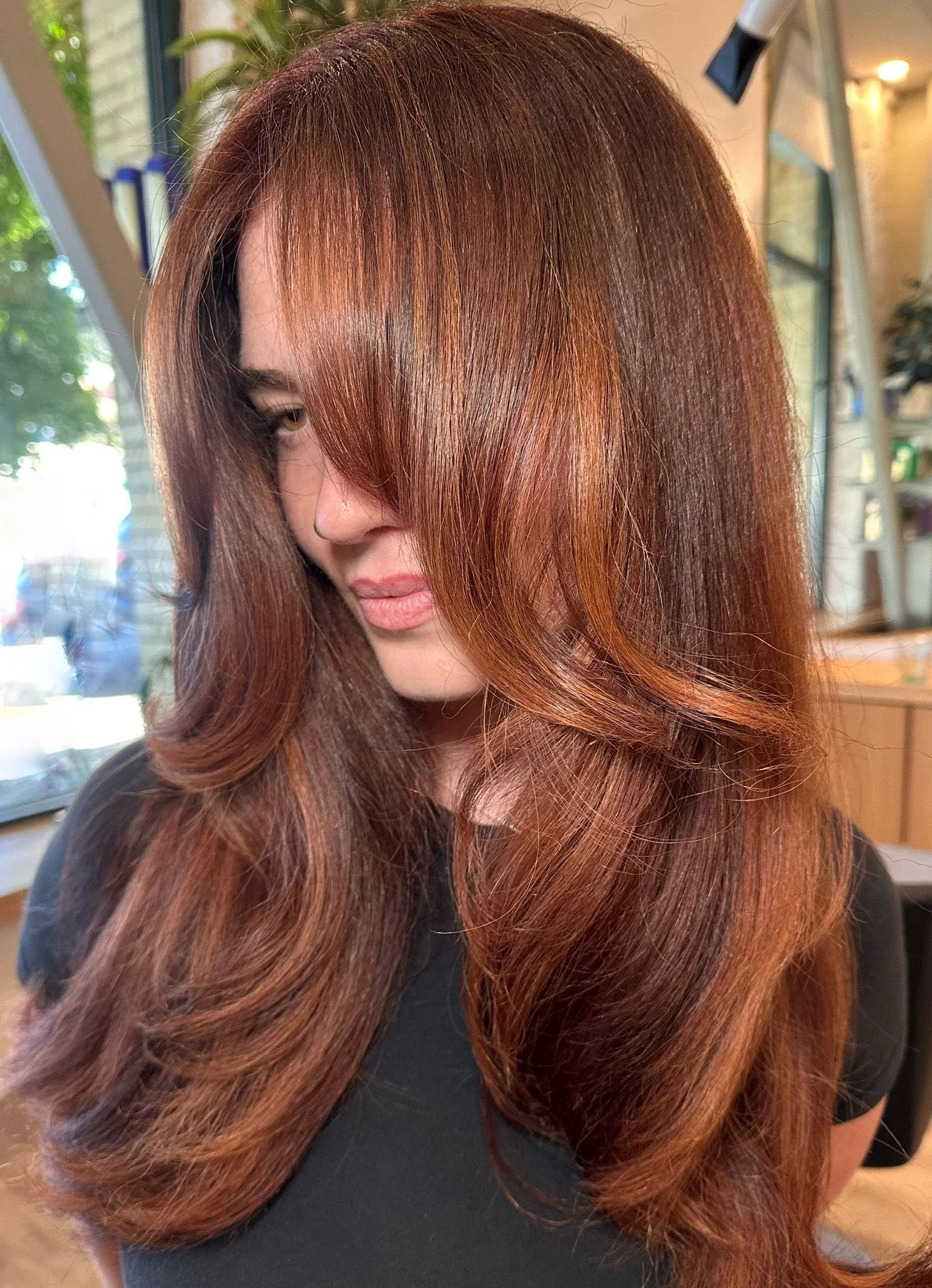 Close-up of a woman with long, shiny, reddish-brown hair styled in soft waves, with a side-swept bang partially covering her face, inside a salon with a large window and salon equipment in the background.