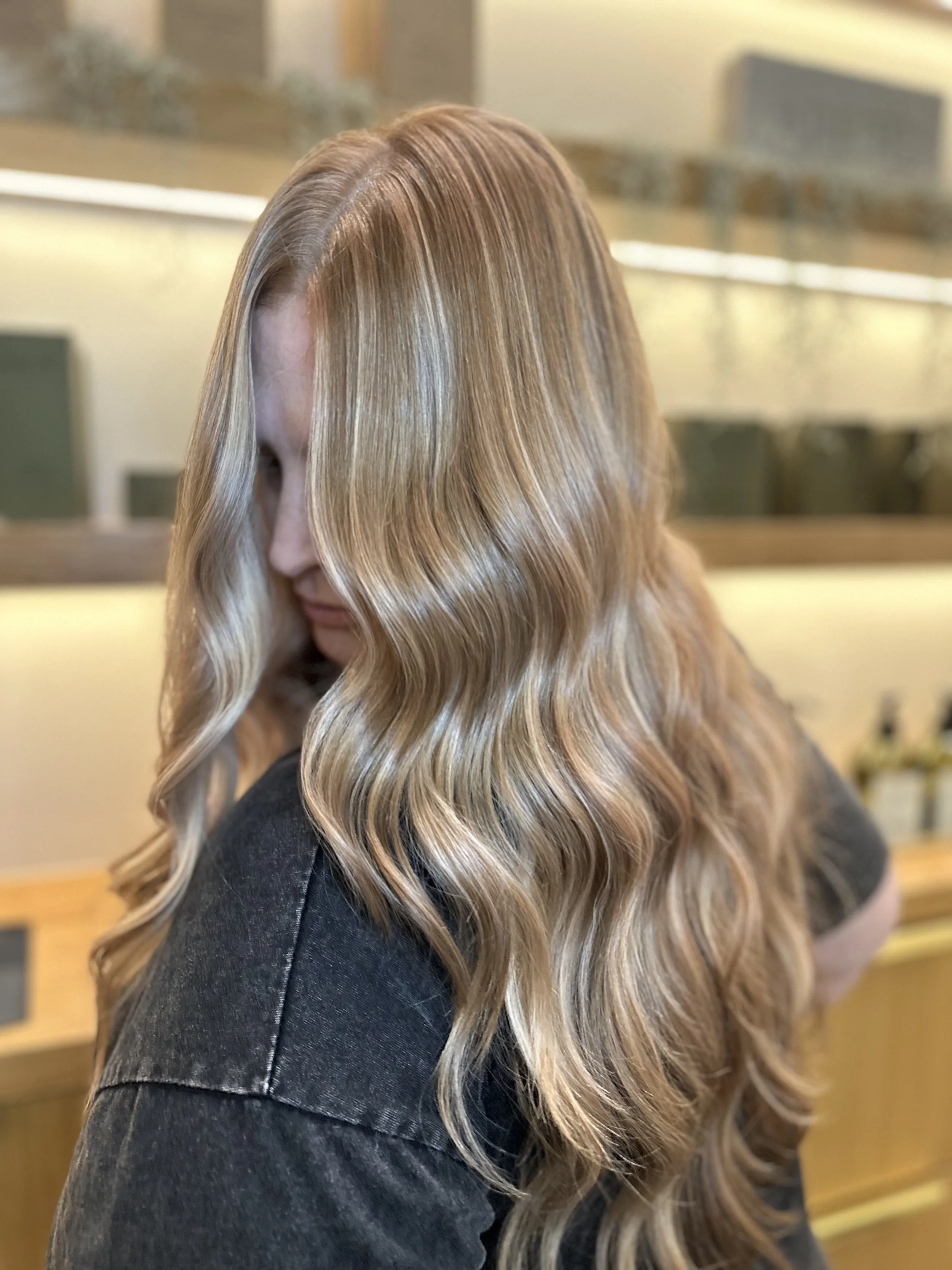Woman with long, wavy blonde hair looking down in a salon or similar setting.
