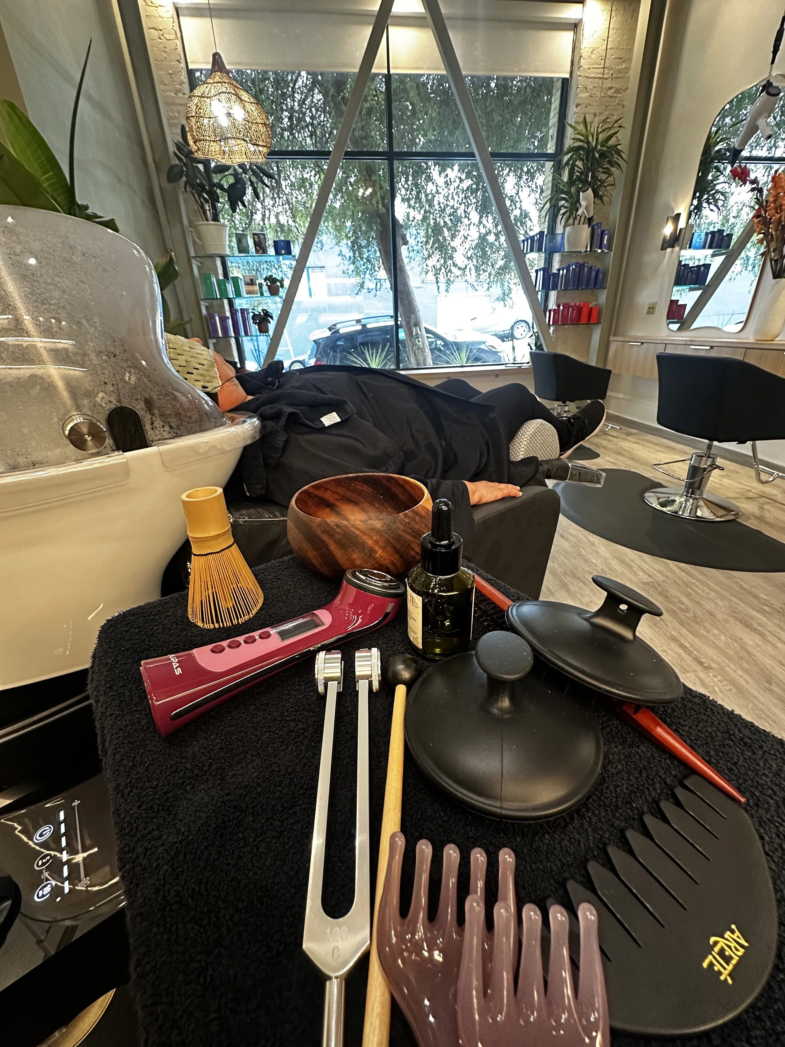 A foot spa salon with a reclining chair, plants, books, and decorative lighting. Tools and cosmetics are on a black towel-covered surface, including a drain board, massage rollers, and bottles.