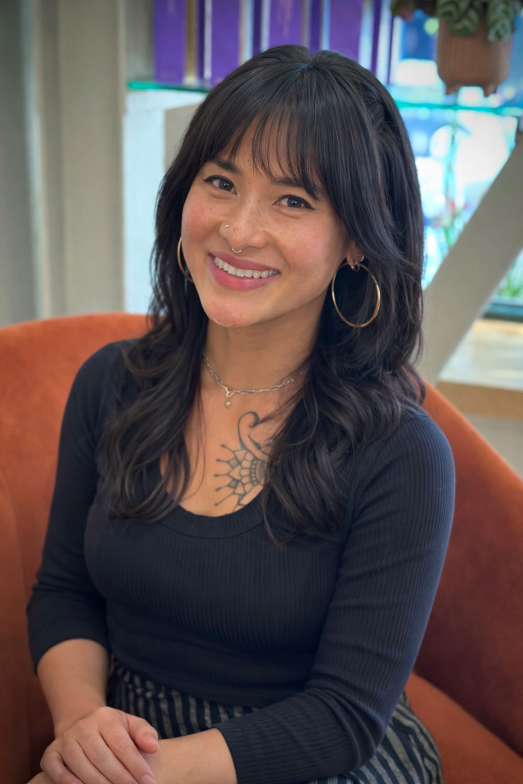 A woman with dark hair, wearing large hoop earrings and a black top, sitting on an orange seat in a cafe, smiling at the camera.