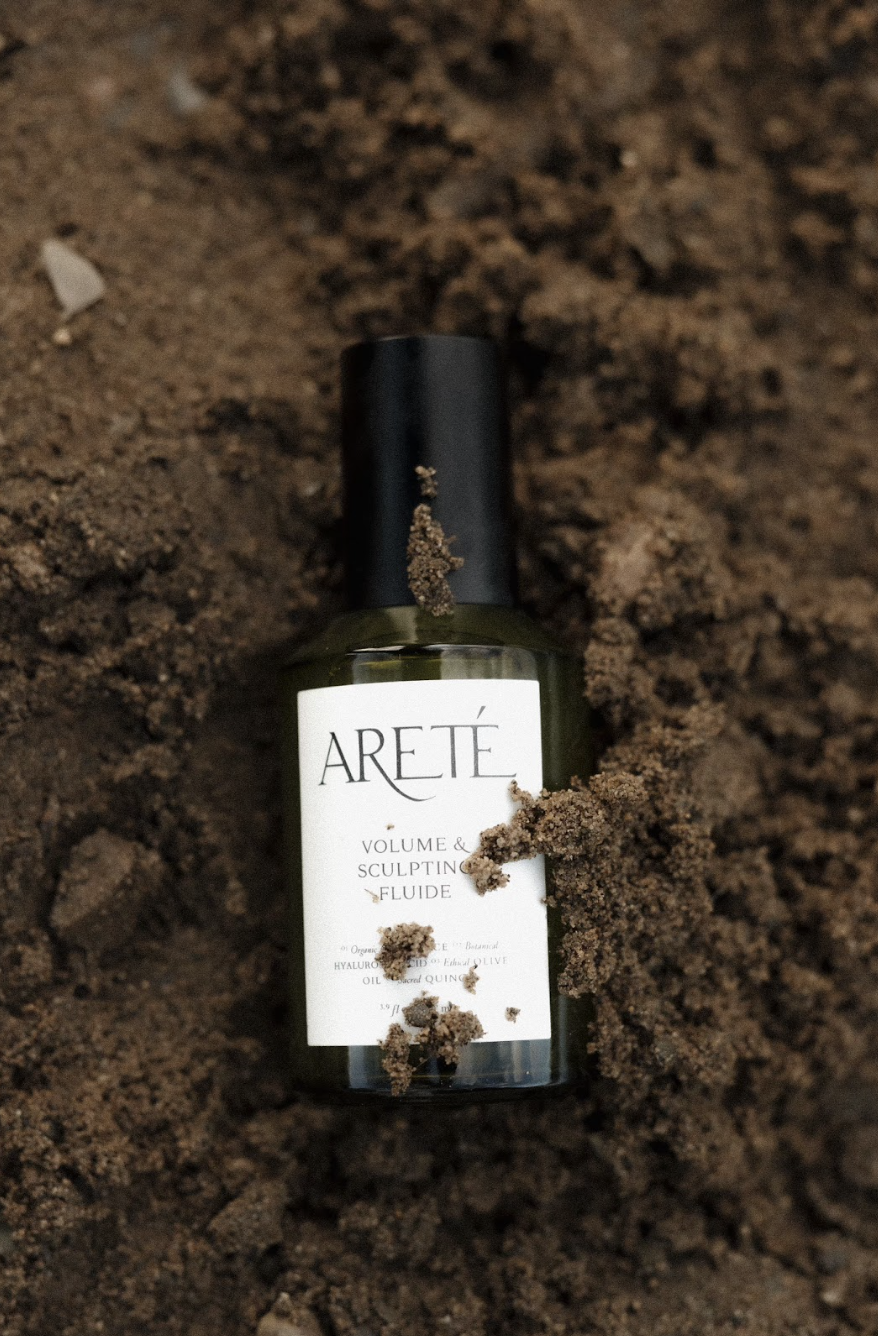 A bottle of ARETÉ Volume & Sculpting Fluid lying on dark soil with some soil particles on the bottle and around it.