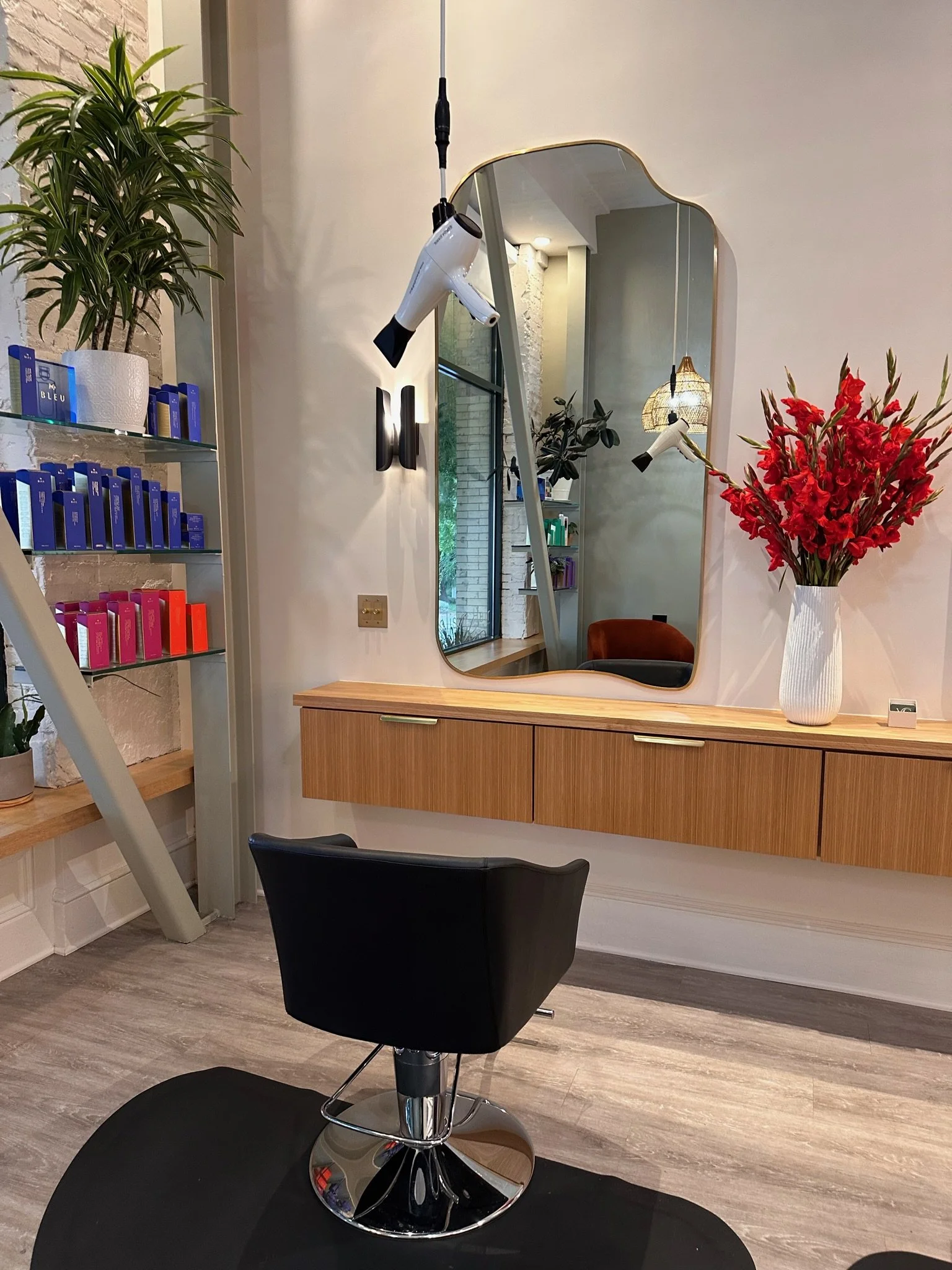 An interior hair salon scene with a black styling chair on a black mat, a wooden counter with closed drawers, a decorative mirror, a tall white vase with red and pink flowers, a potted plant, a shelf with hair products, and a large mirror reflecting the room.