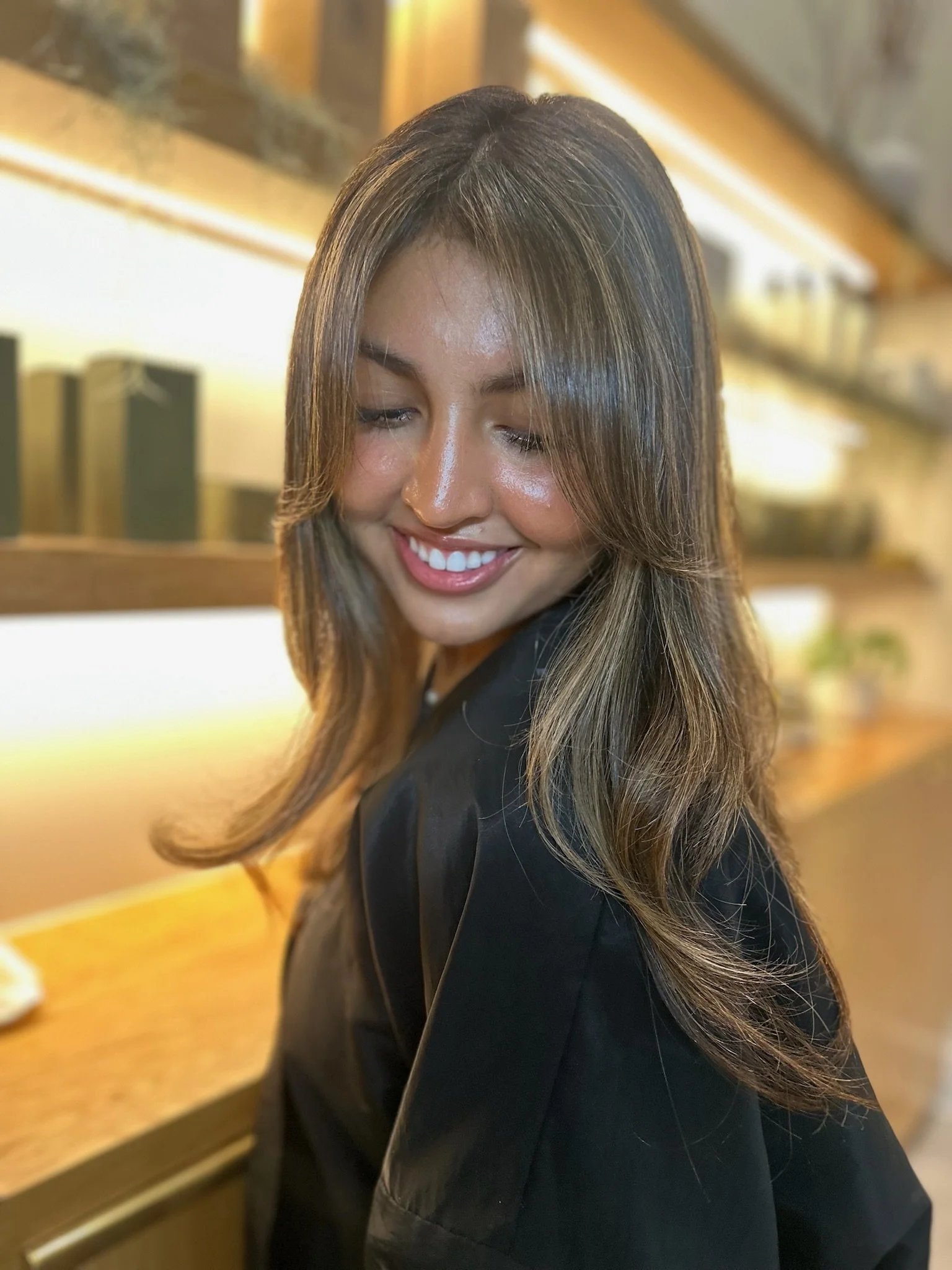 Smiling woman with long brown hair in an indoor setting with warm lighting.