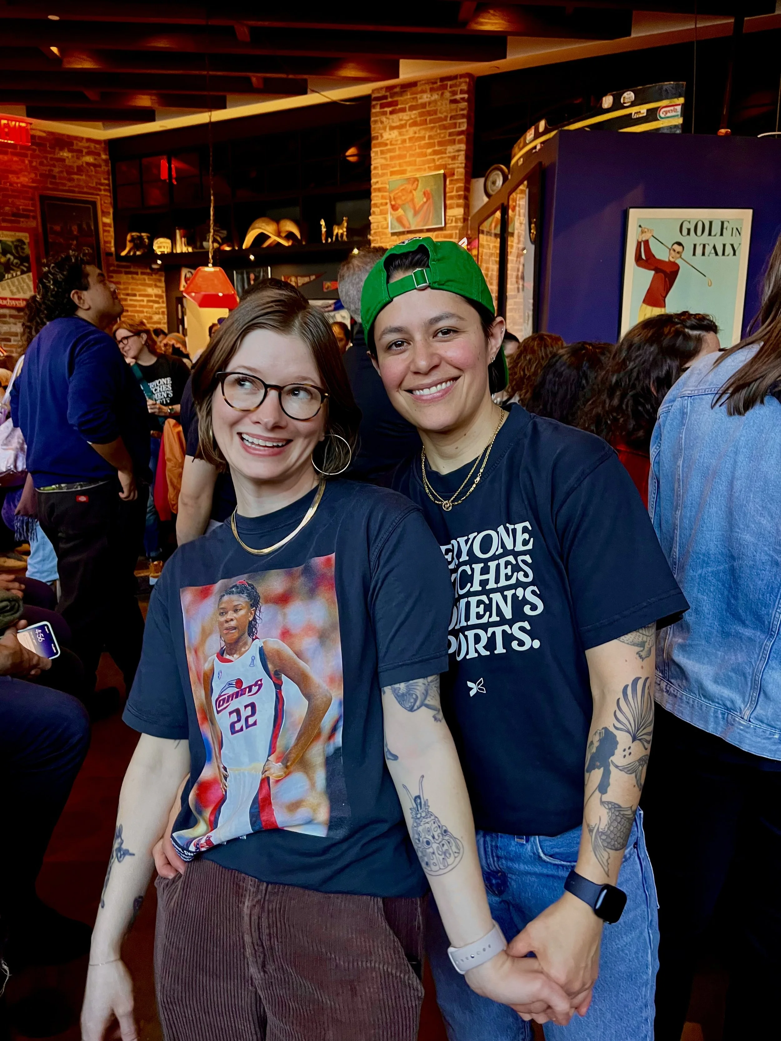 Athena Keke's Bar owners pictured, two women smiling and holding hands at a crowded indoor event with brick walls and vintage posters, wearing dark t-shirts and showing their tattoos.