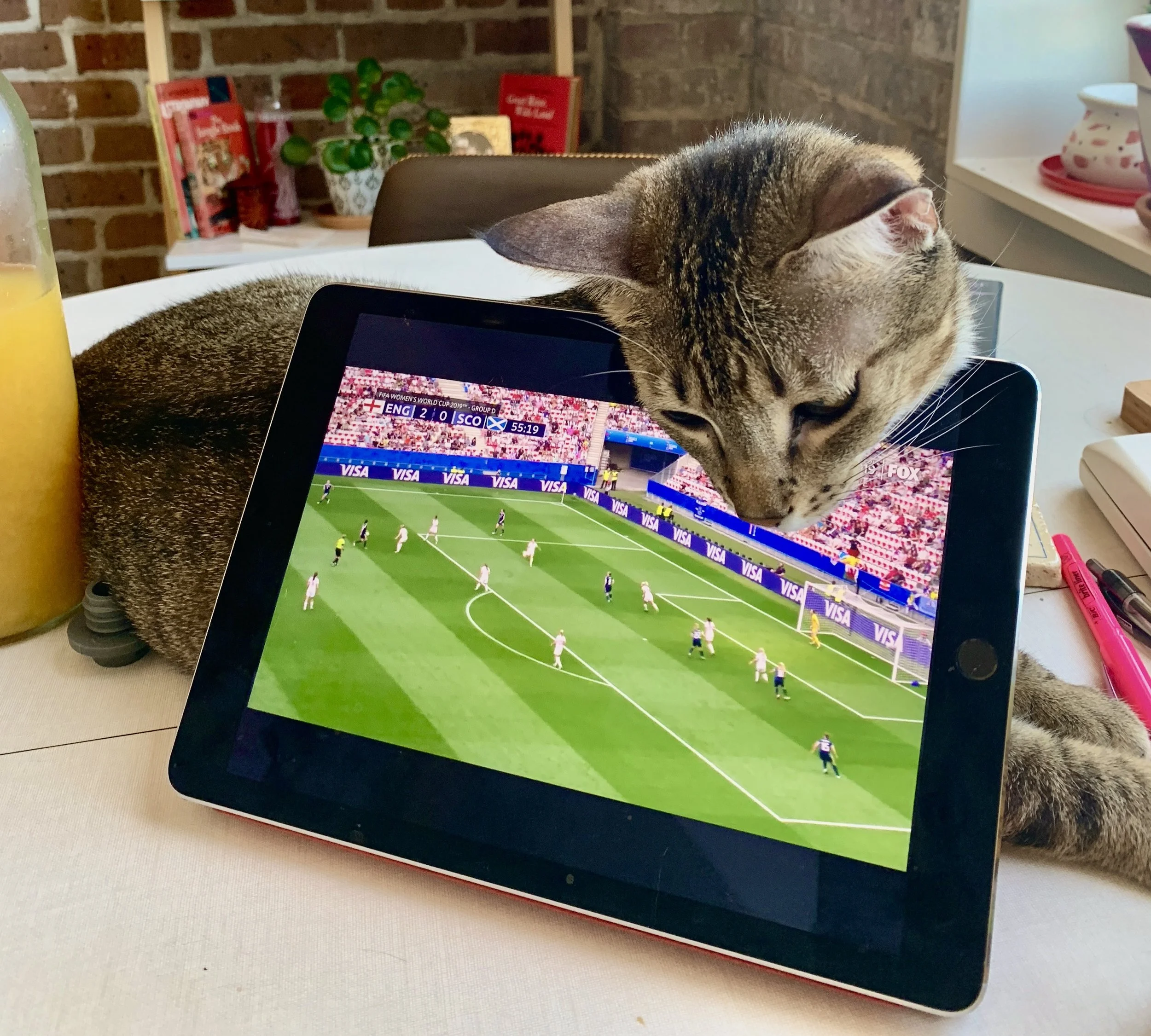 A tabby cat peering over a tablet displaying a soccer game, with a glass of orange juice and some pens on a table.