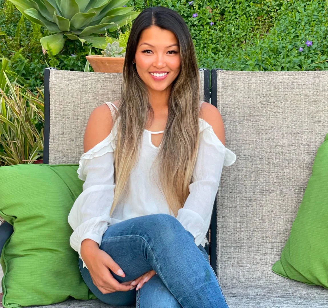 Woman smiling on an outdoor chair with green cushions, surrounded by plants and greenery.