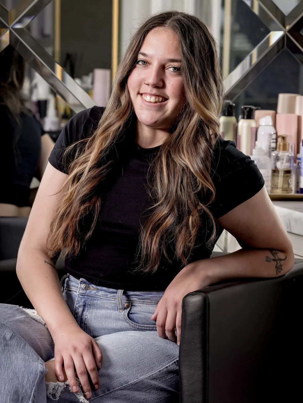 A woman with long hair sitting on a chair, wearing a black shirt and jeans, smiling, with beauty products visible in the background.